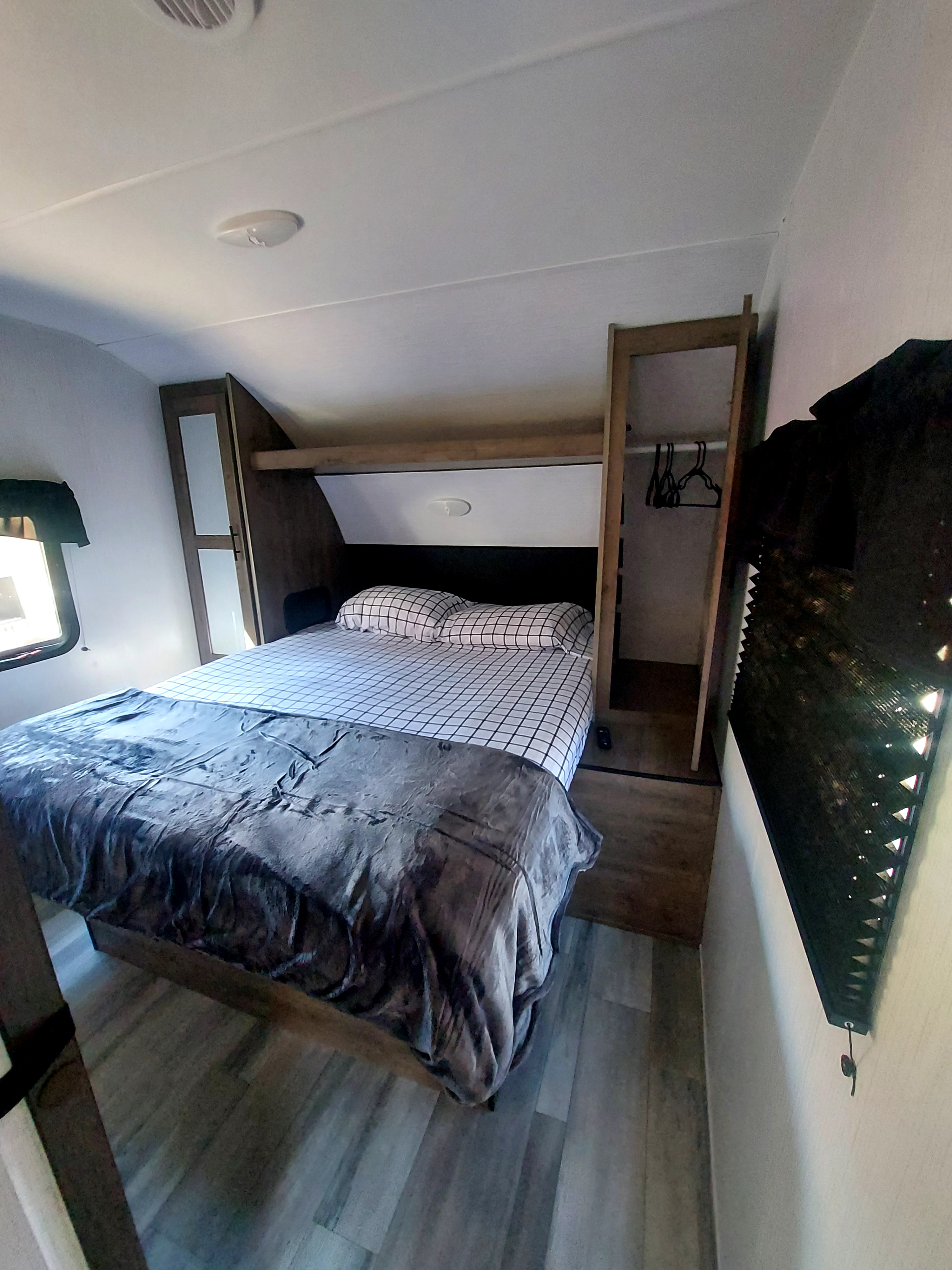 This RV rental bedroom has a bed with grid sheets, a dark blanket, wood storage, and an open closet. Ideal for travel.. Heartland RV Pioneer 2023