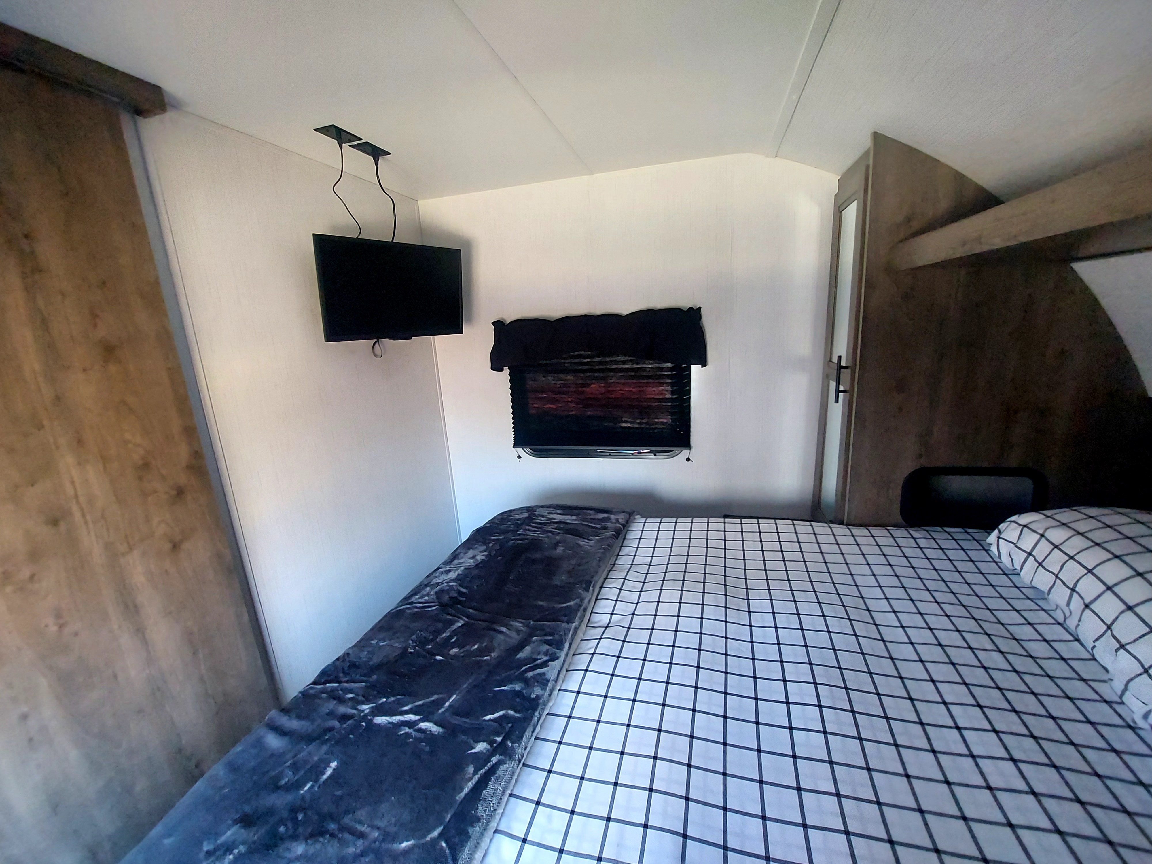 Cozy **rv rental** bedroom featuring a checker-patterned bed, wall-mounted TV, and window with a shade.. Heartland RV Pioneer 2023
