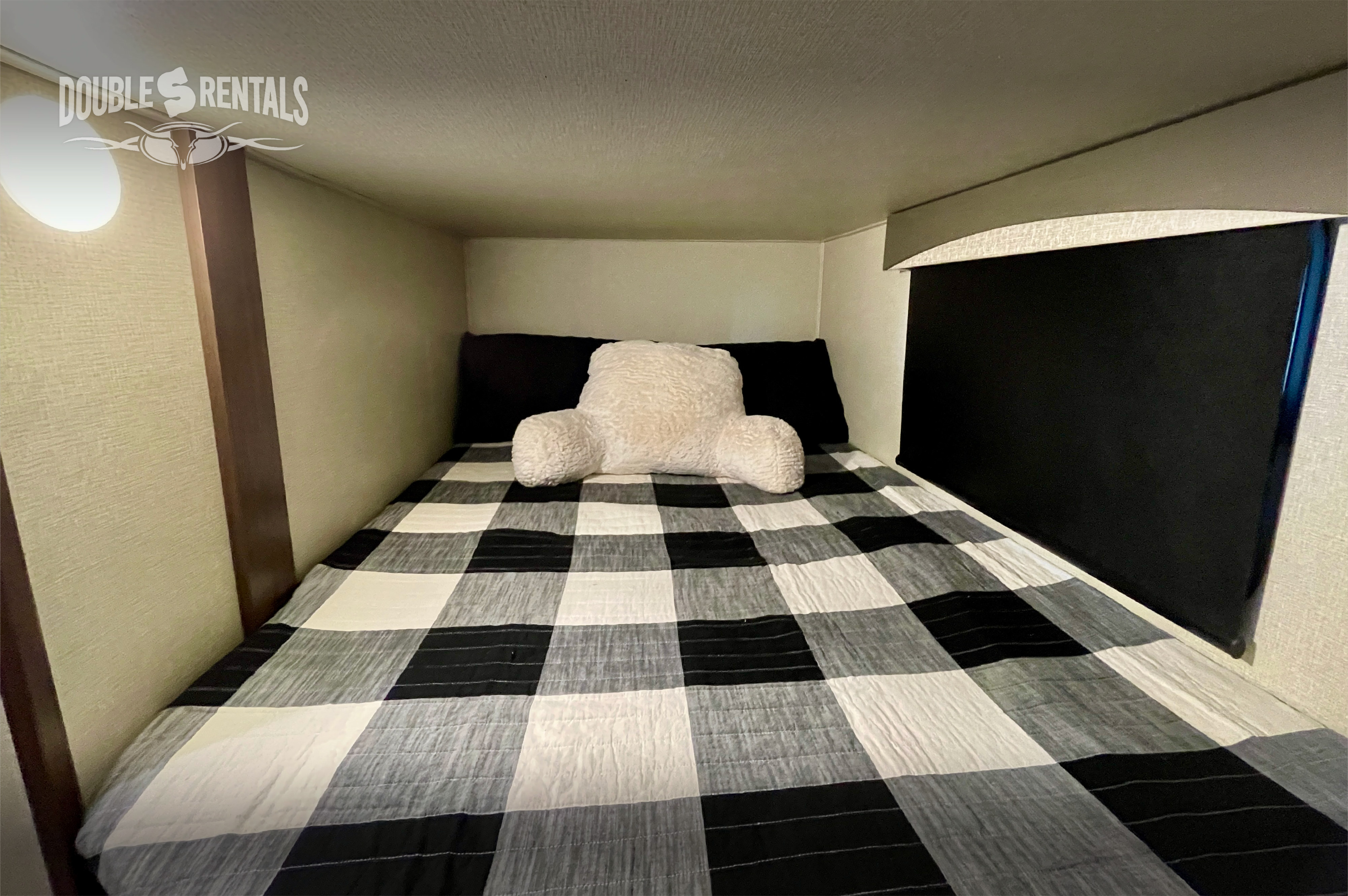 Cozy RV rental bunk bed features a black & white buffalo check comforter, a white backrest pillow, and a reading light.. Prime Time Crusader Lite 2020
