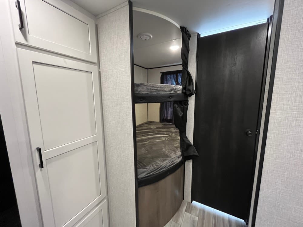 Inside an RV rental: white storage cabinets, a bunk bed area with black curtains, and a dark door leading elsewhere.. Jayco Jay Flight 2024