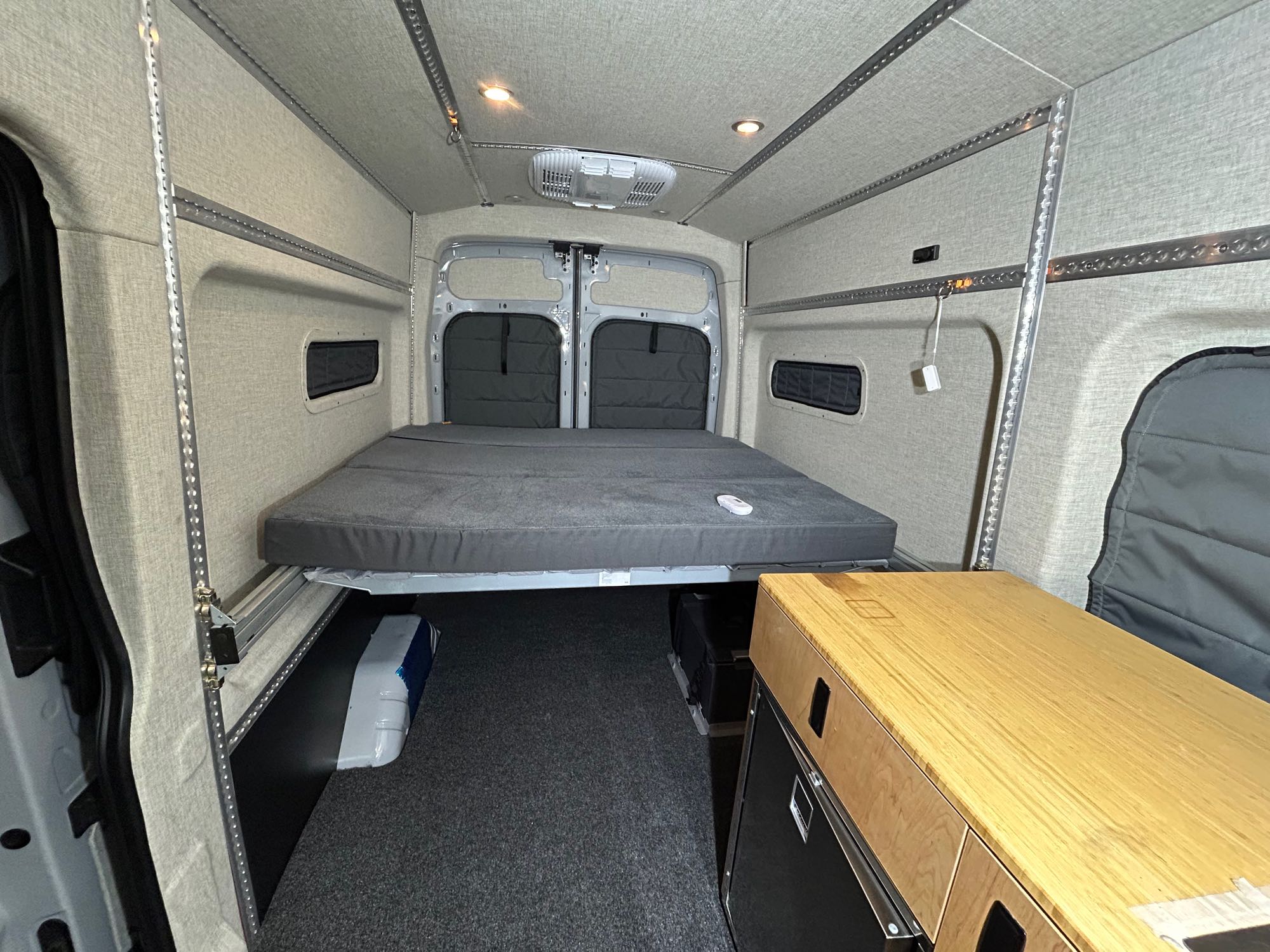 RV rental: Van interior with a raised bed, under-bed storage, and walls lined with light-colored fabric and mounting tracks.. Ford Transit 2023