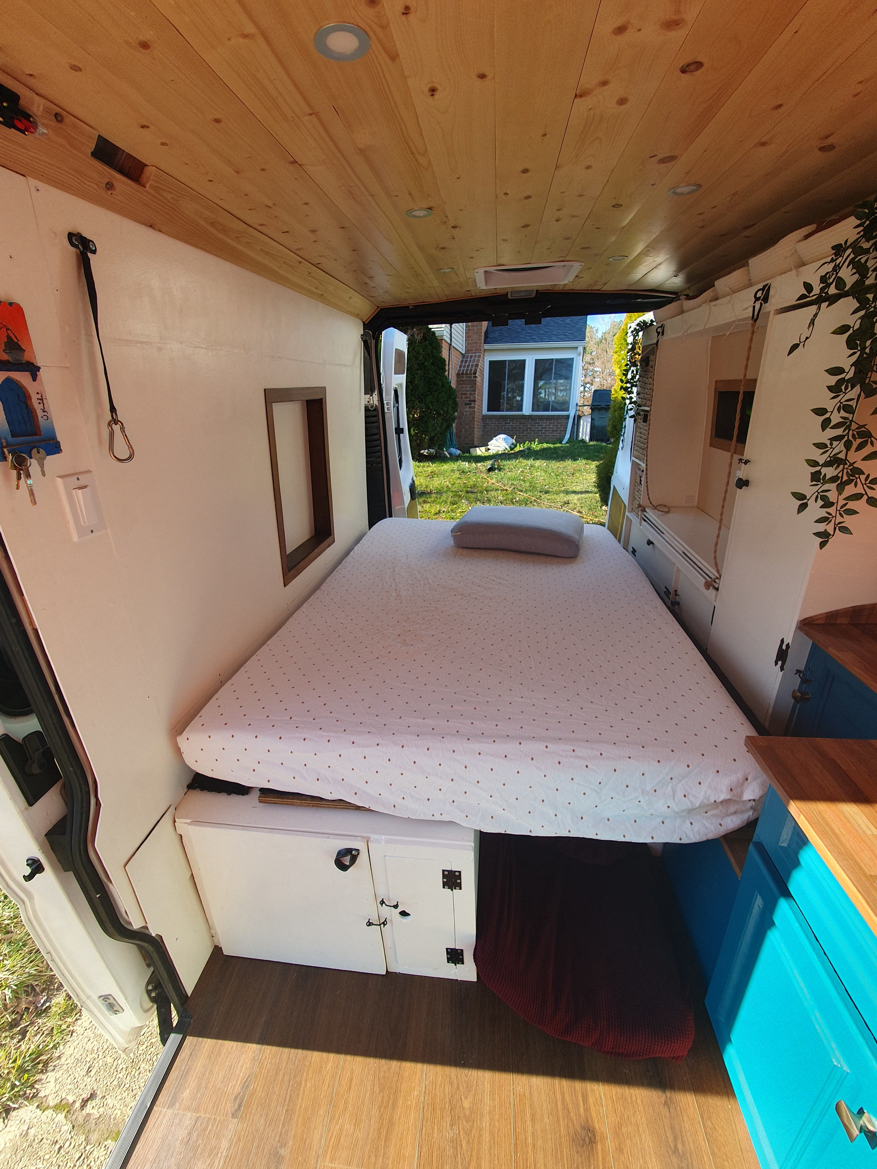 Bright camper van interior for your rv rental: comfy bed, wood ceiling, and custom storage. Open doors reveal a residential backyard.. Dodge Sprinter Van 2016