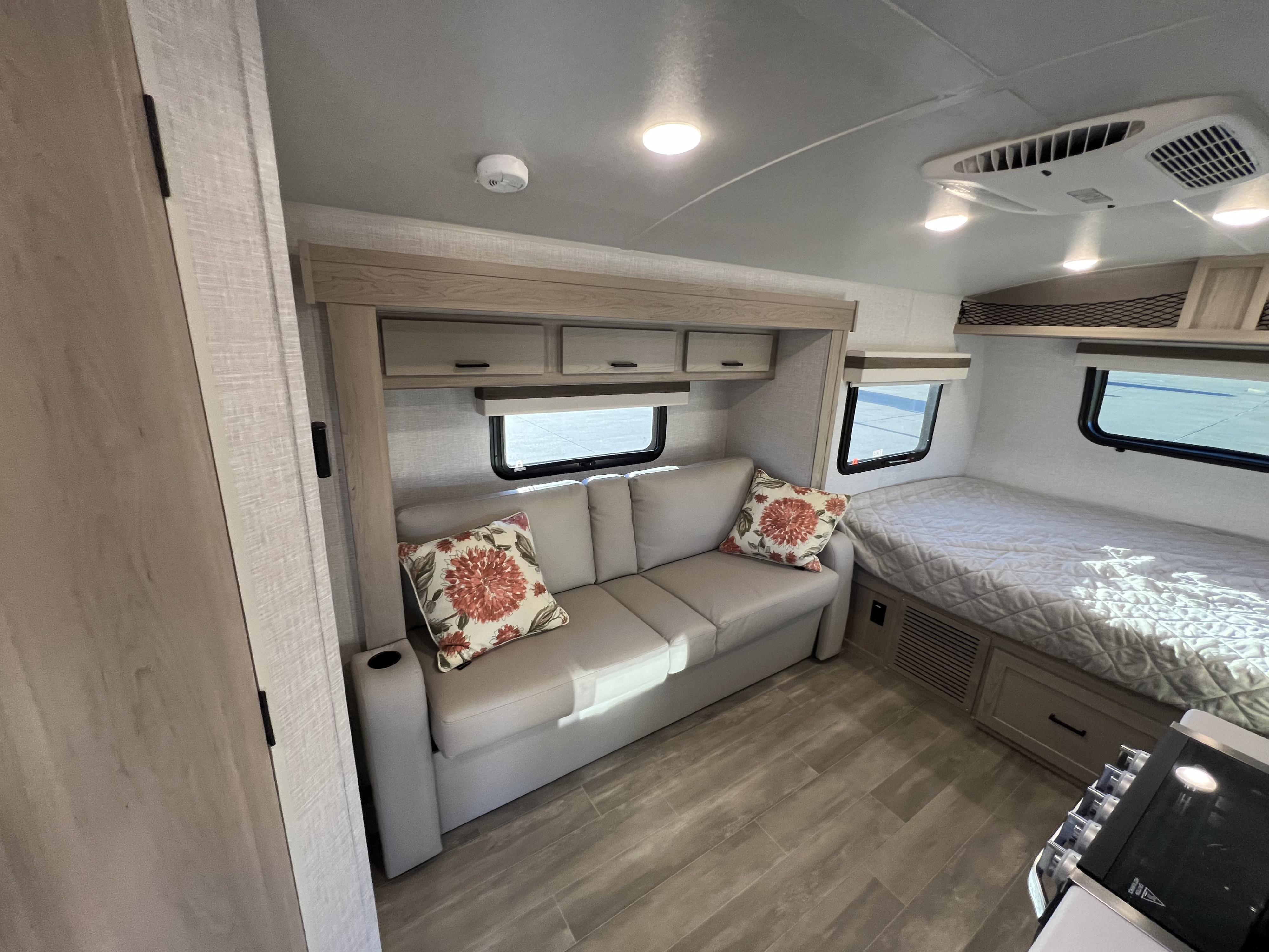Modern RV rental interior showcasing a neutral-toned sofa, overhead cabinets, and a tidy bed area with windows.. Forest River Rockwood Geo Pro 2023