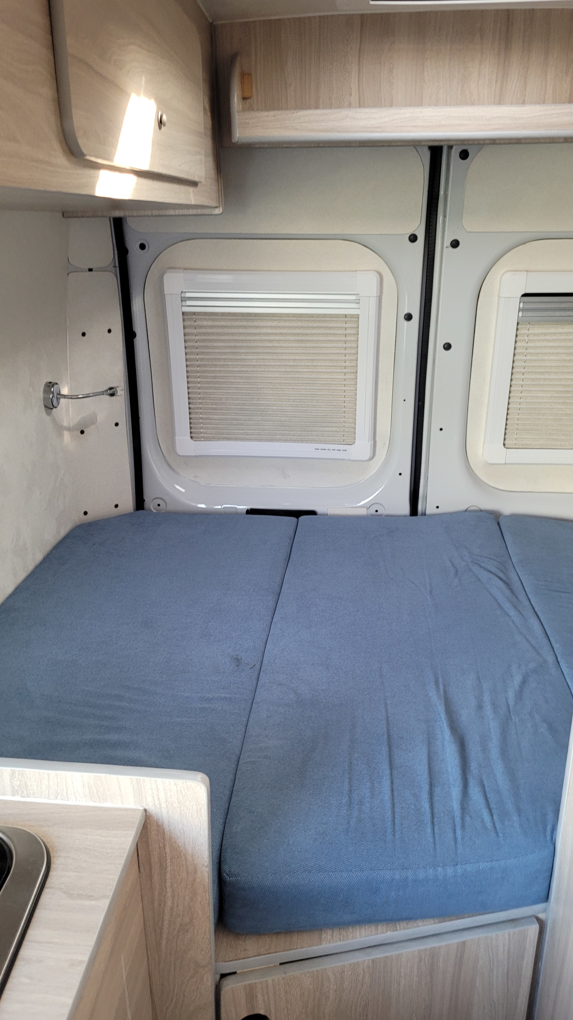 Inside an RV rental: a neat sleeping area with a blue mattress, overhead storage, and two windows with blinds.. Fiat Ducato 2018