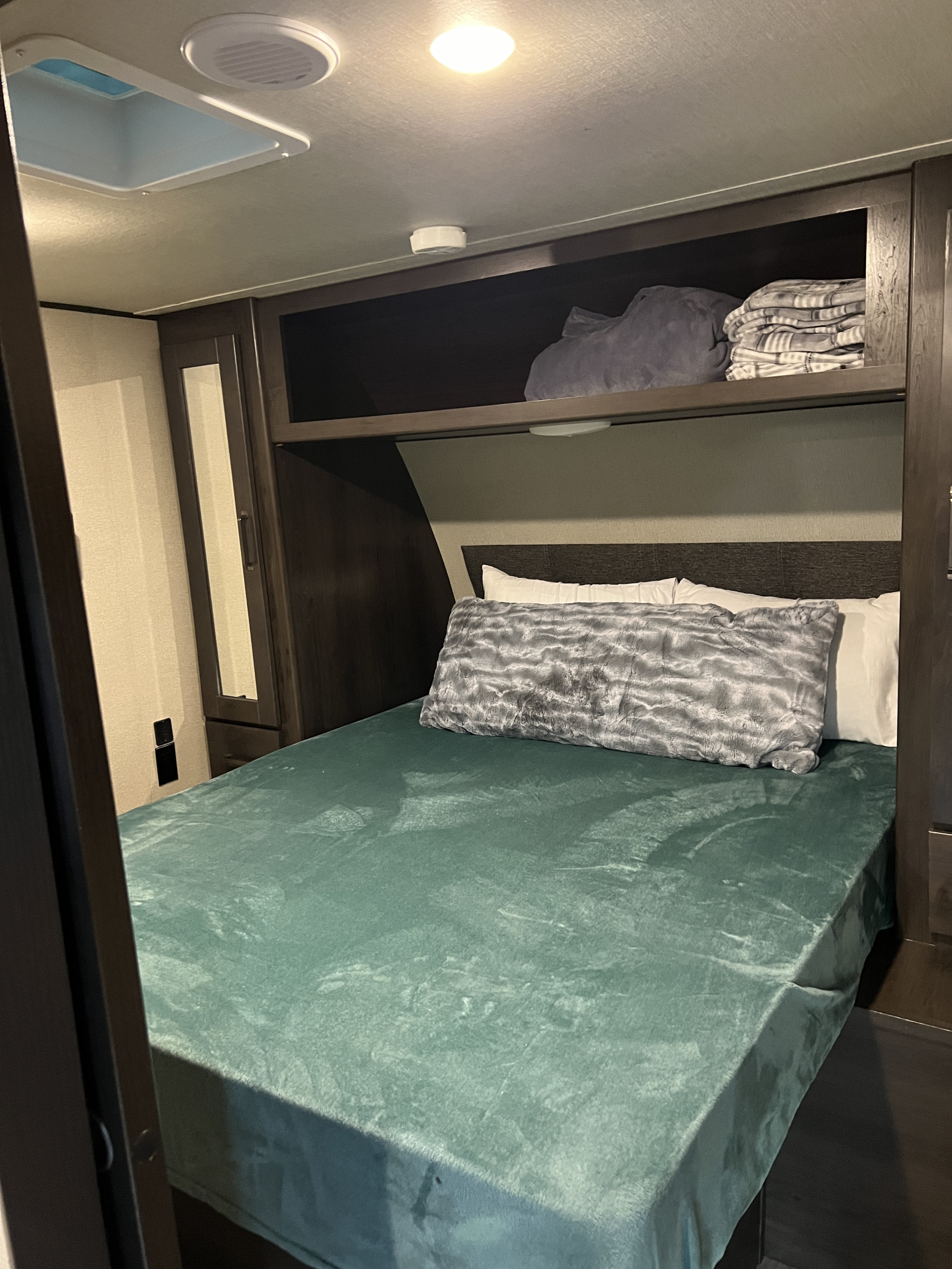 Cozy RV rental bedroom featuring a teal bedspread, overhead storage, and ceiling vent, providing a comfortable space.. Grand Design Transcend Xplor 2023