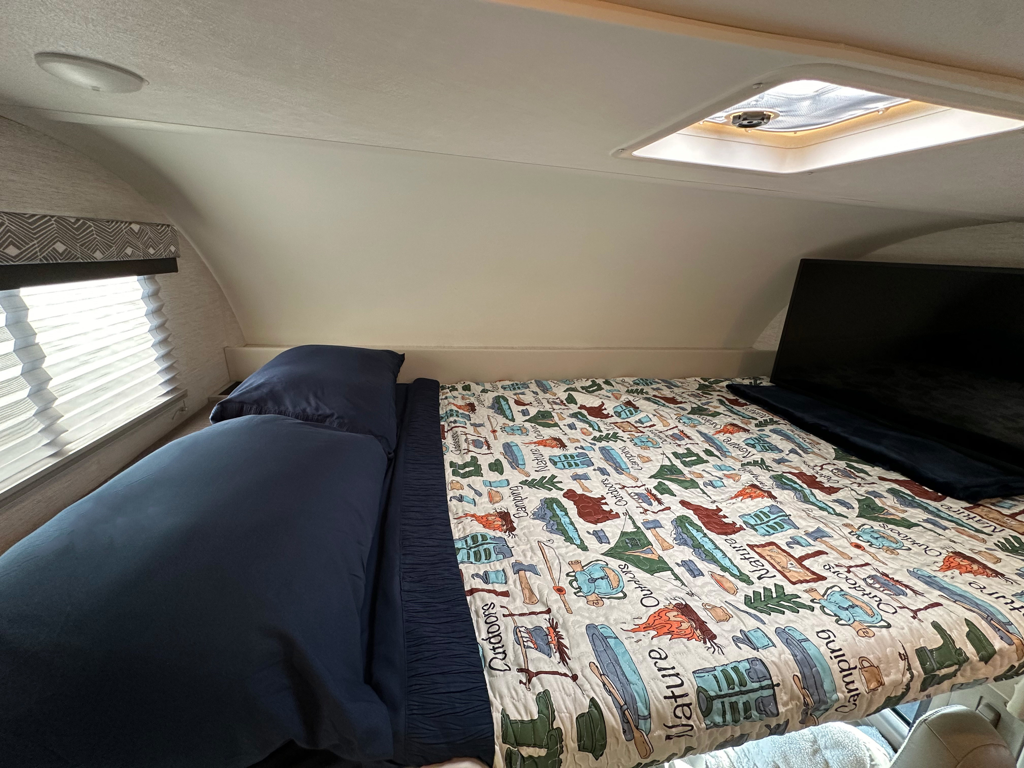 Cozy RV rental sleeping area featuring a camping-themed quilt, window blinds, and skylight for your adventure.. Thor Four Winds 2024