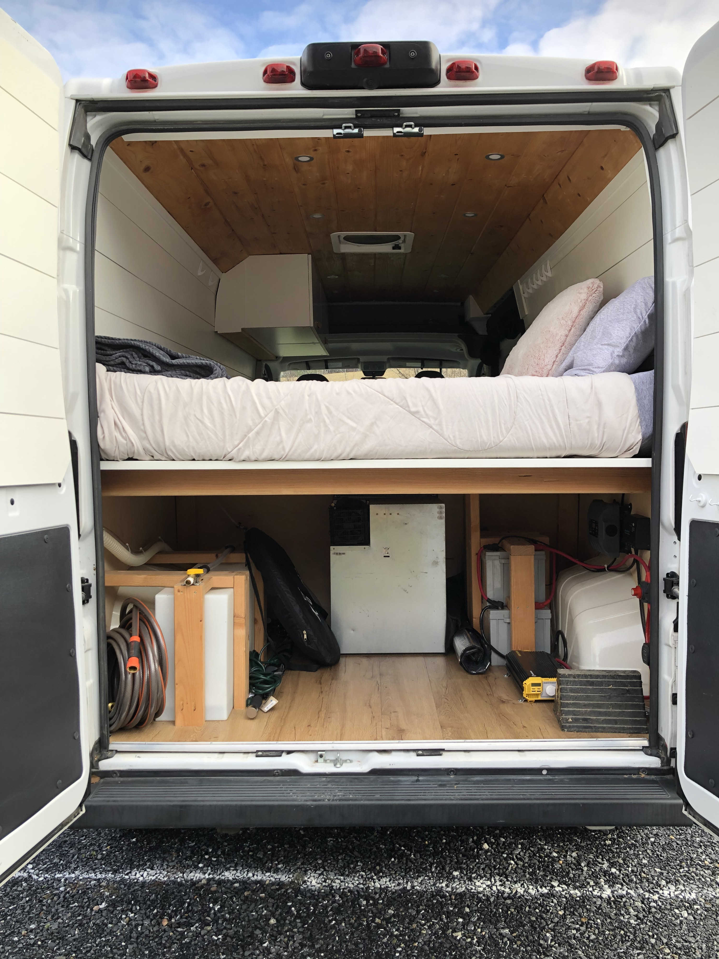 Inside an RV rental: comfy bed, wood ceiling, and under-bed storage with fridge, batteries, & water tanks.. Dodge Promaster 2500 2021