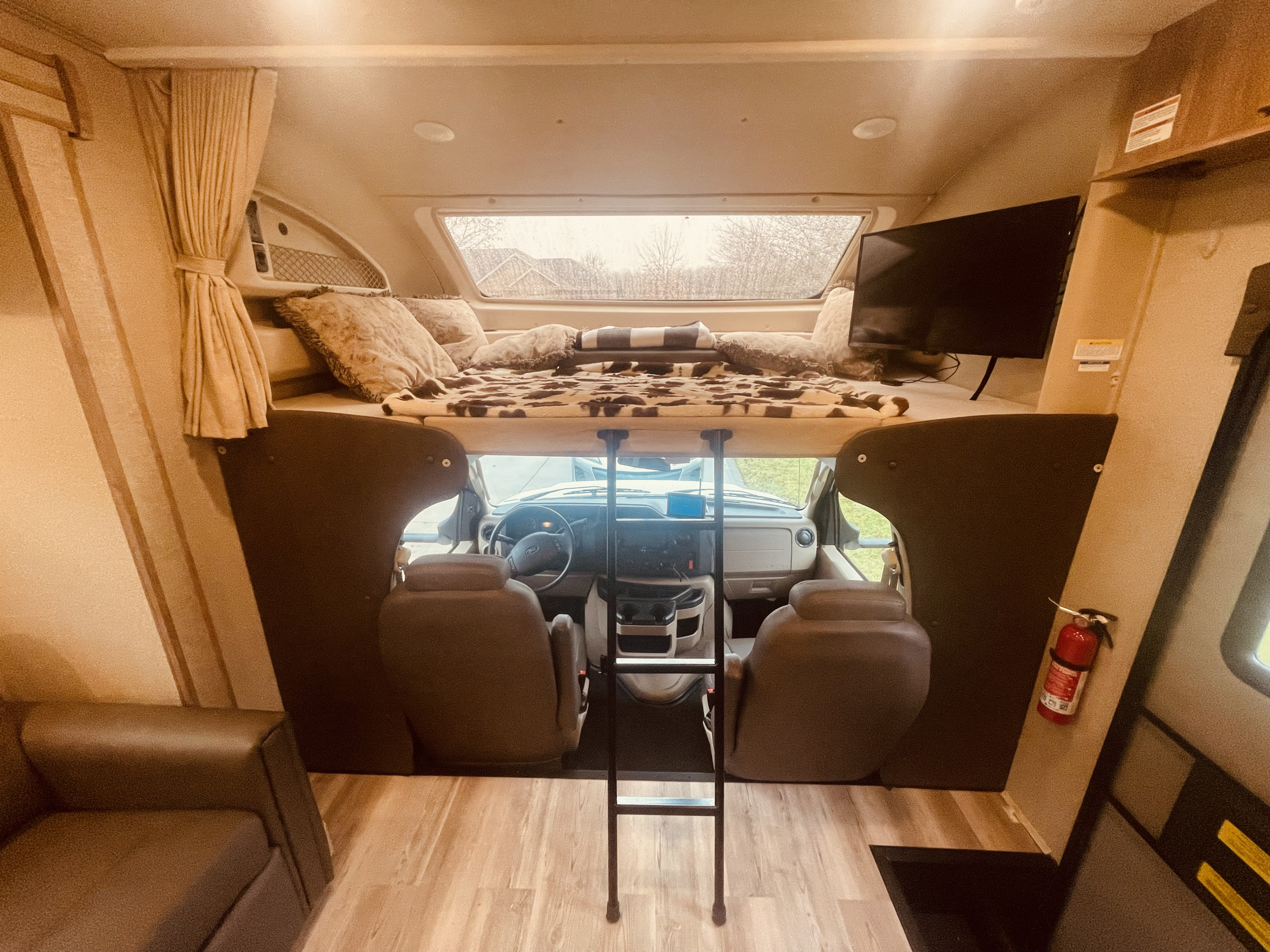 Cozy RV rental interior: overhead bed with cow print blanket, TV, ladder, and front driving area visible.. Entegra Coach Odyssey 2020