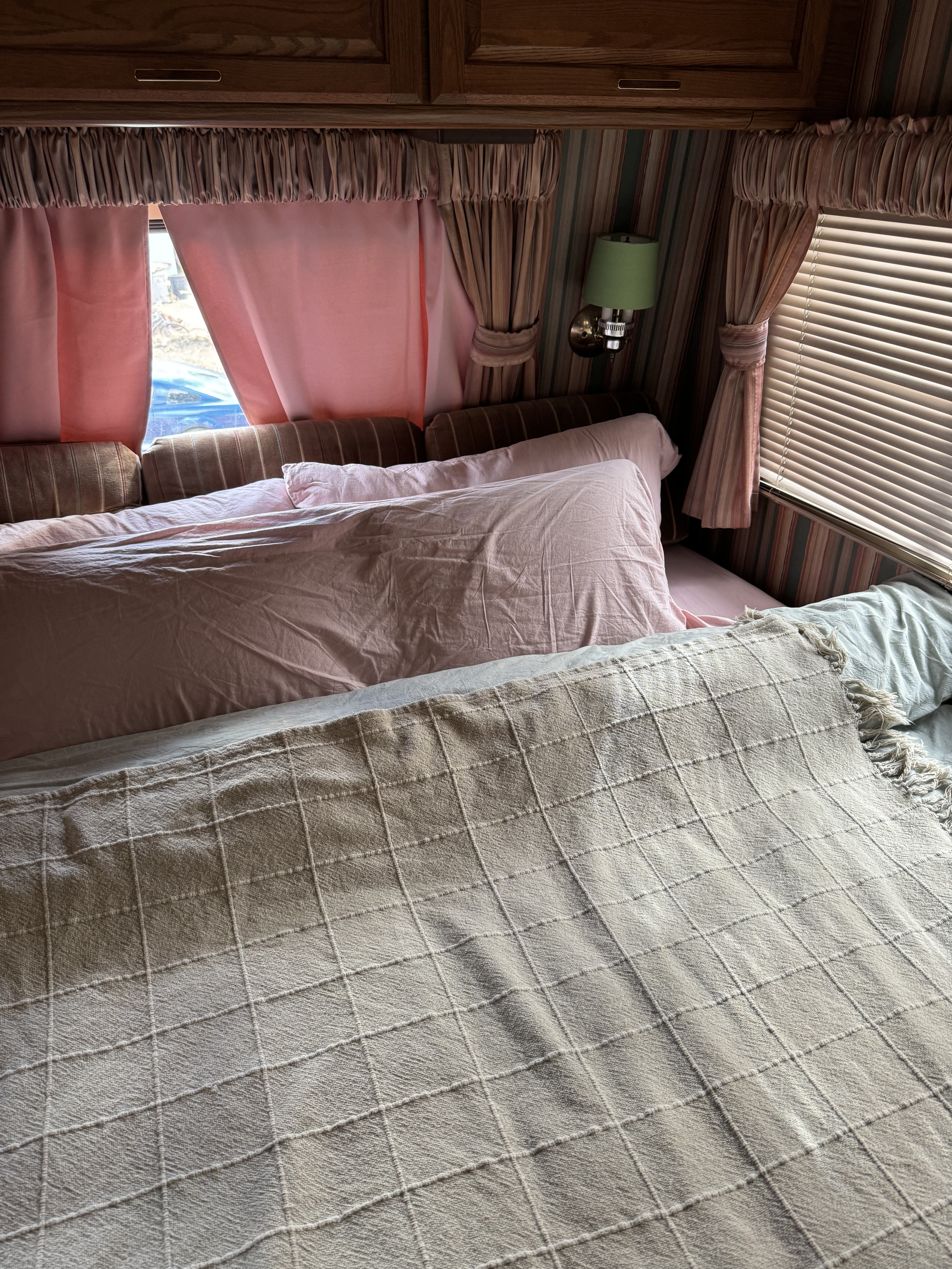 Inside an RV rental: a cozy bedroom with pink sheets, a checkered blanket, and vintage wood paneling and curtains.. Winnebago Chieftain 1986