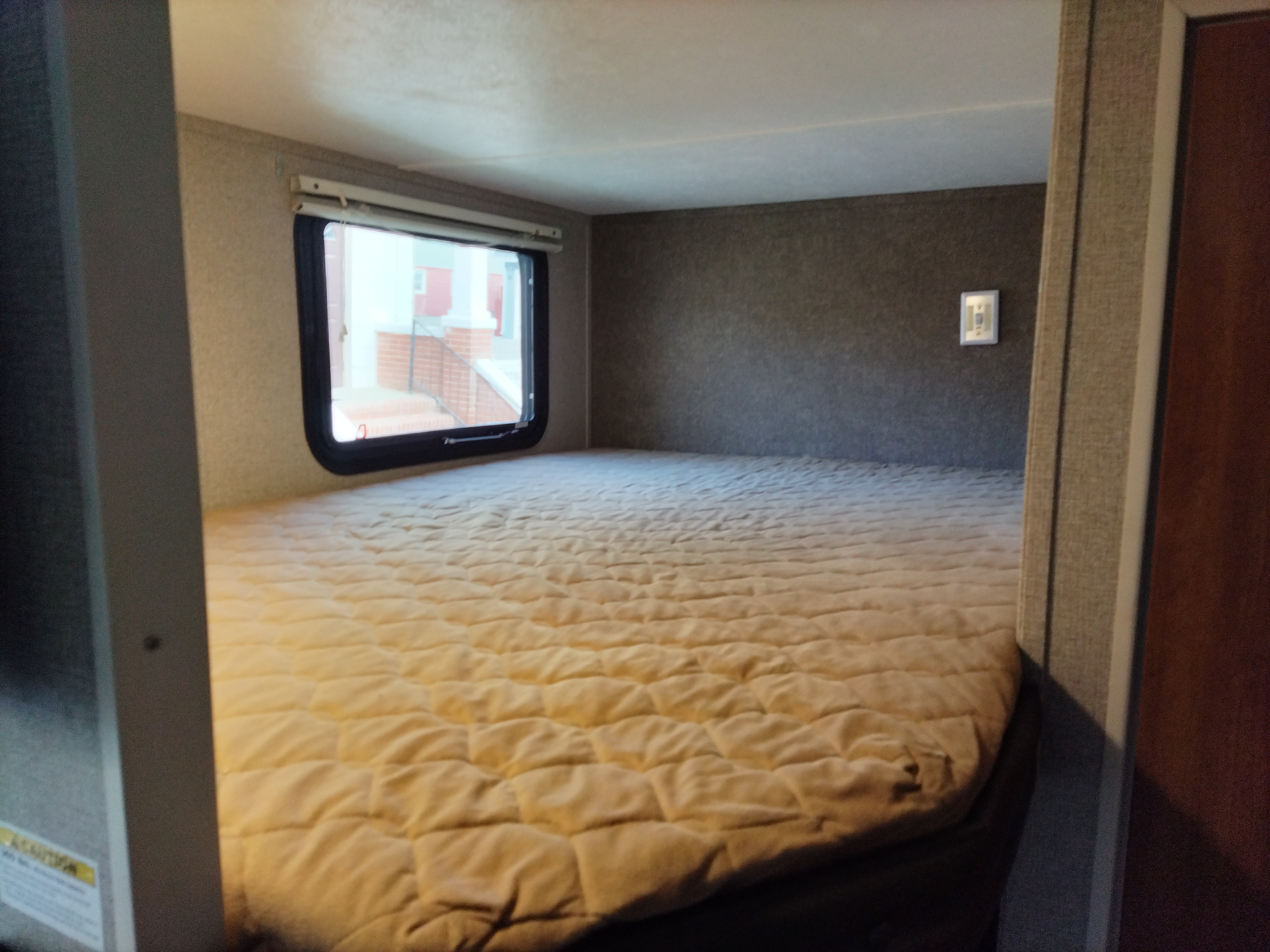 This RV rental features a wide, light-colored quilted bed by a window. Outside, a brick house with steps is visible.. Keystone RV Springdale Eco-Lite 2018