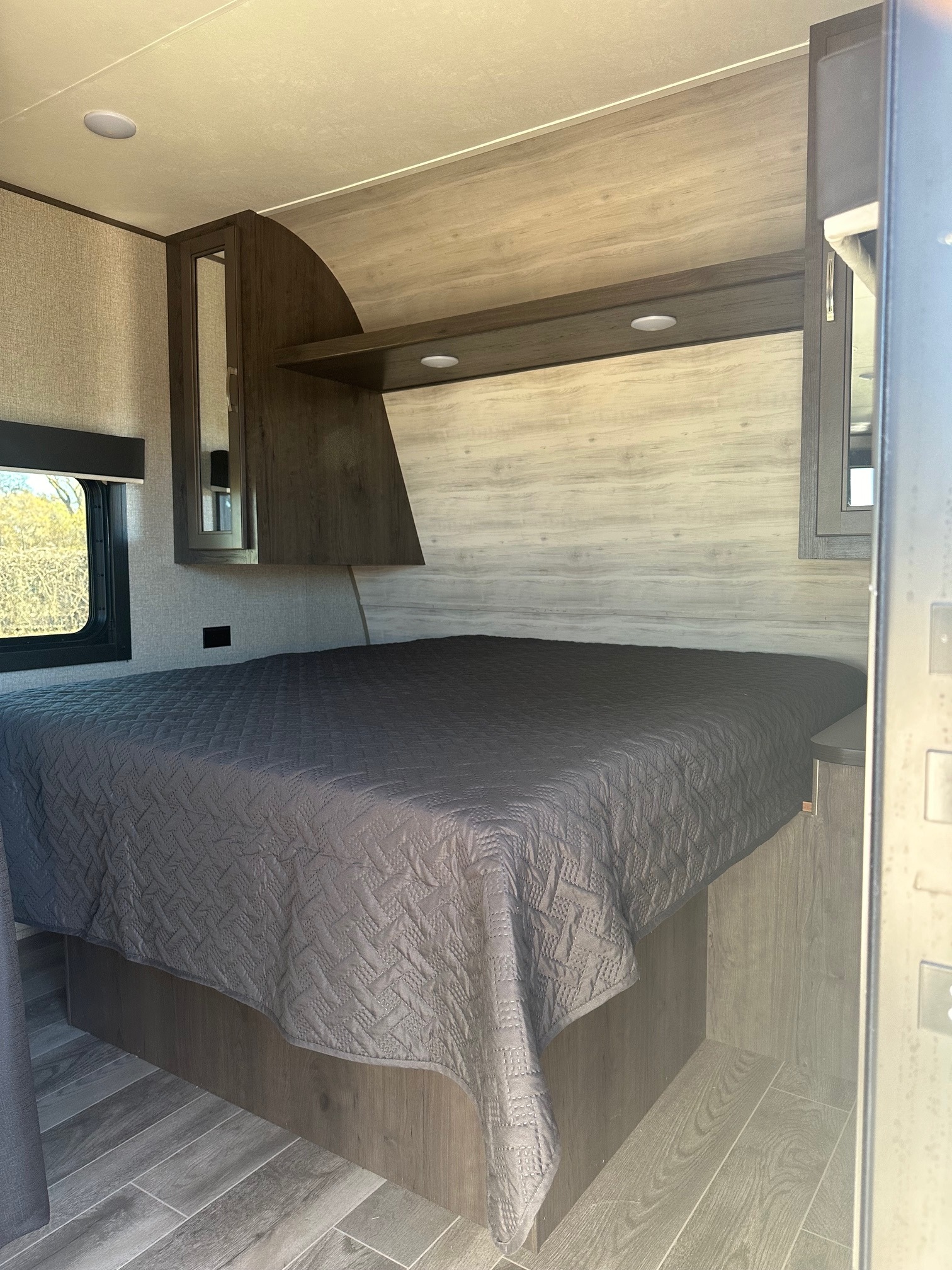 This RV rental features a comfortable bedroom with a dark gray quilted bed, wood paneling, overhead cabinets, window, and lighting.. Jayco Jay Flight 2021
