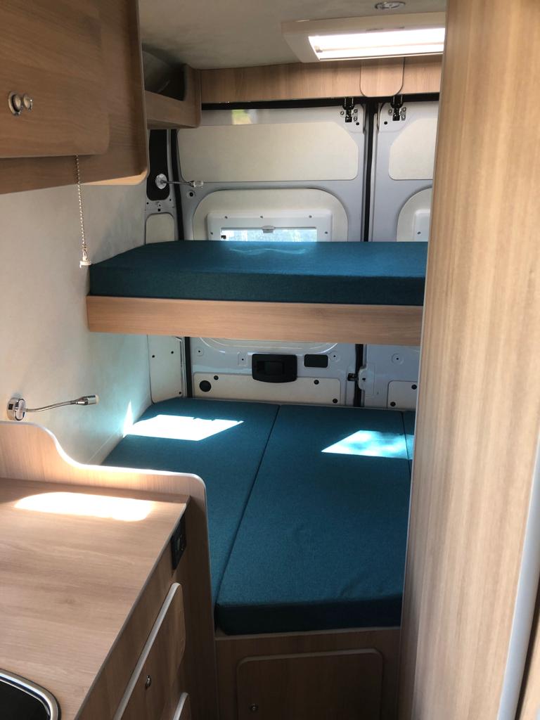 Inside an RV rental: light wood interior with teal bunk beds. Upper bunk, lower bed/seating, and small kitchen counter.. Fiat Ducato 2018