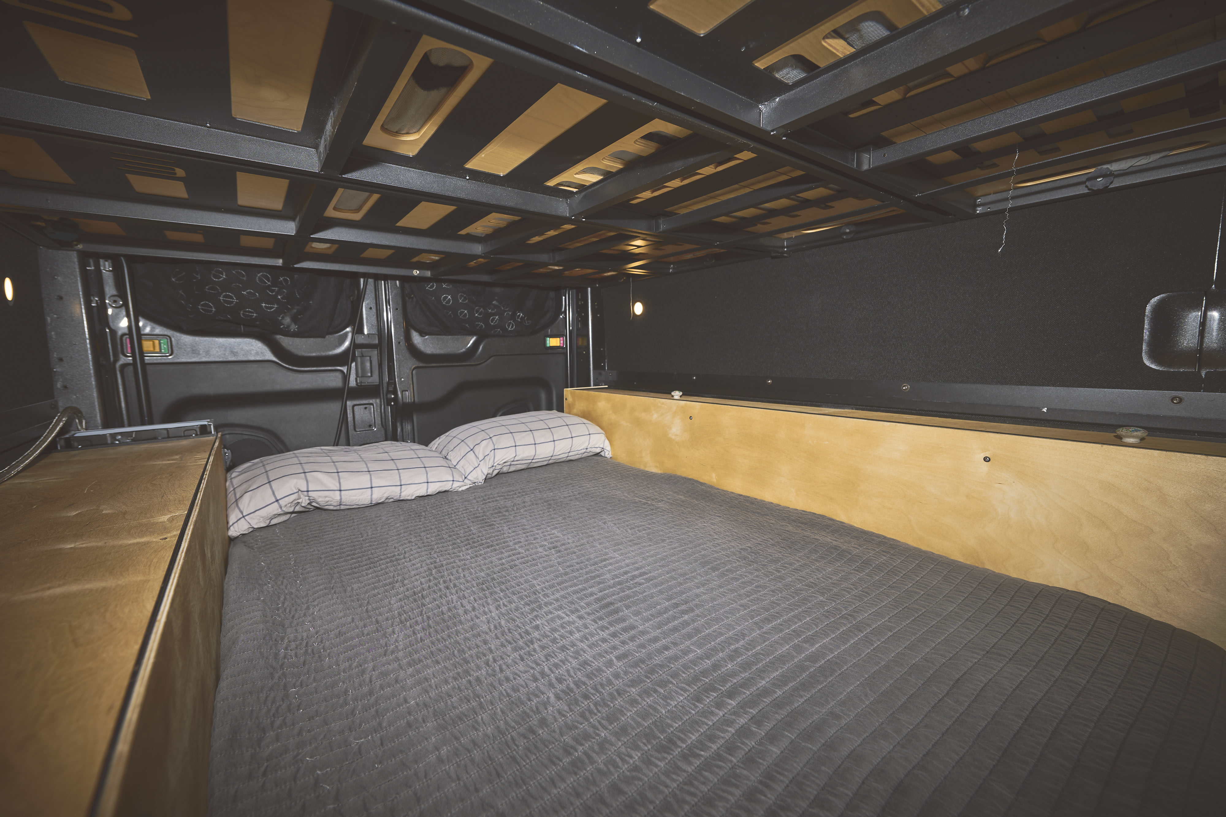 Cozy RV rental bedroom showing a made bed with grey quilted blanket & plaid pillows, light wood cabinetry, and structural overhead storage.. Ford Transit 2020