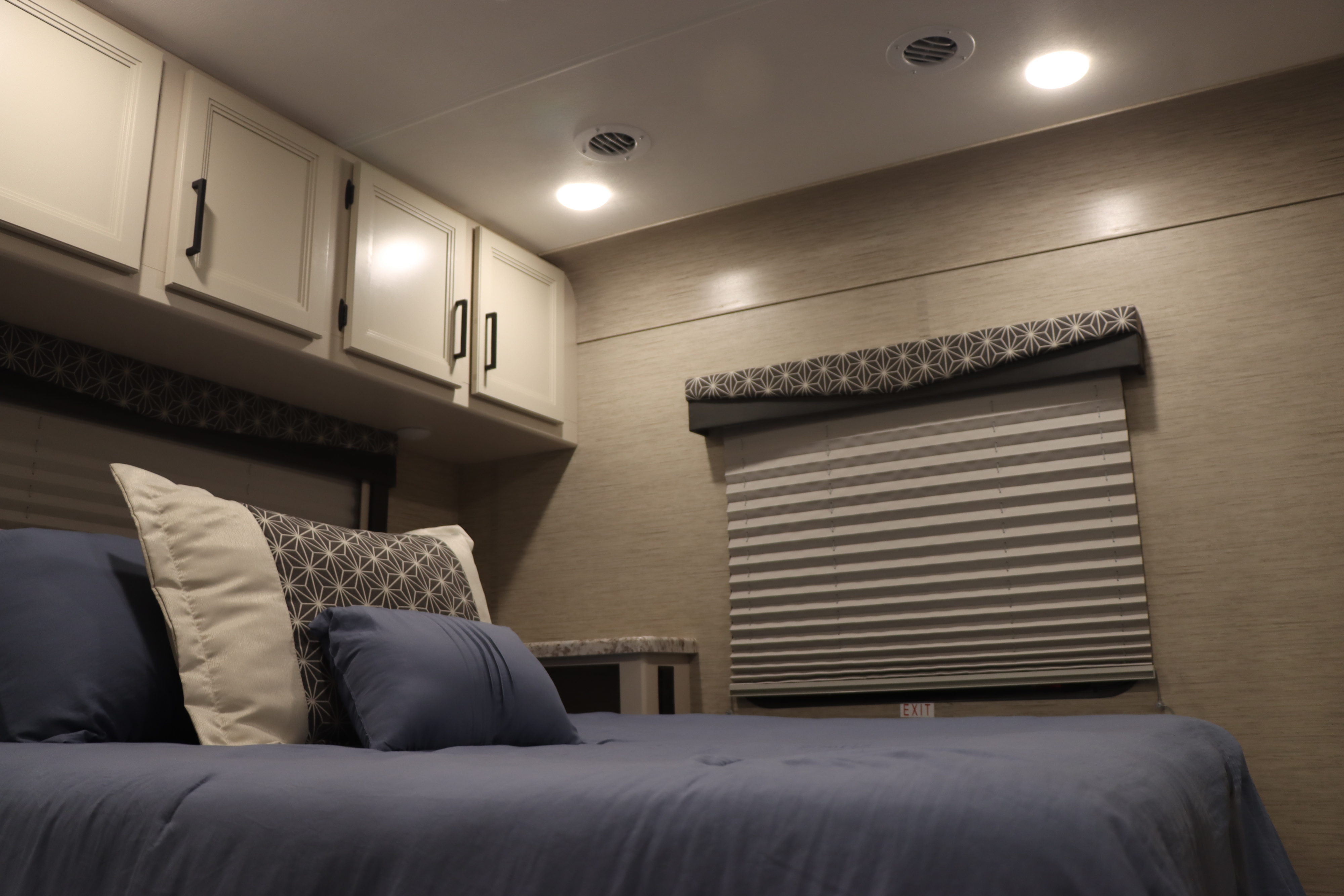 Cozy RV rental bedroom with blue bedding, bright lights, white cabinets, and patterned window treatments.. Thor Freedom Elite 2023