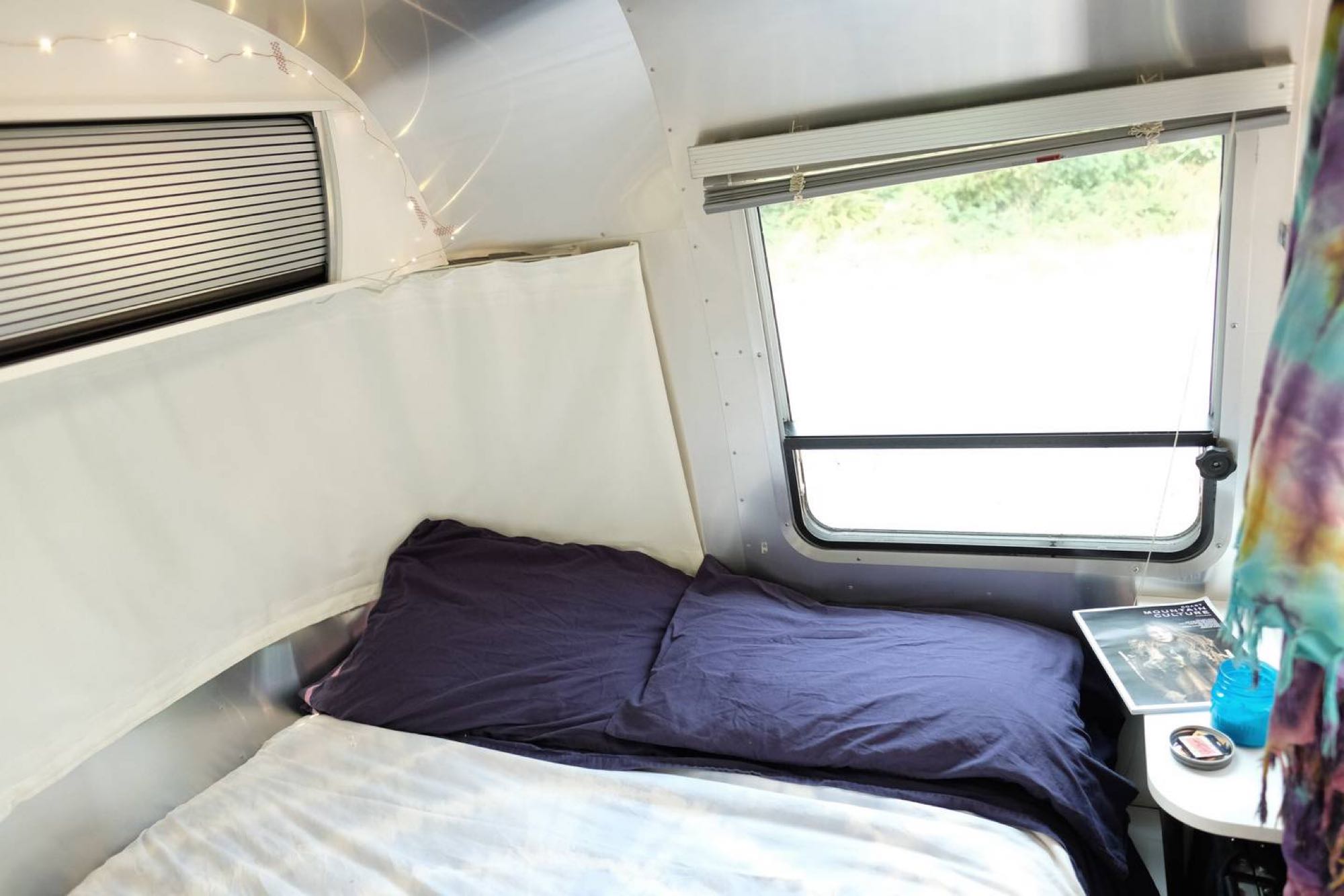 Experience a cozy RV rental bedroom: bed with purple pillows, metallic walls, ambient string lights, and windows.. Airstream Sport 2016