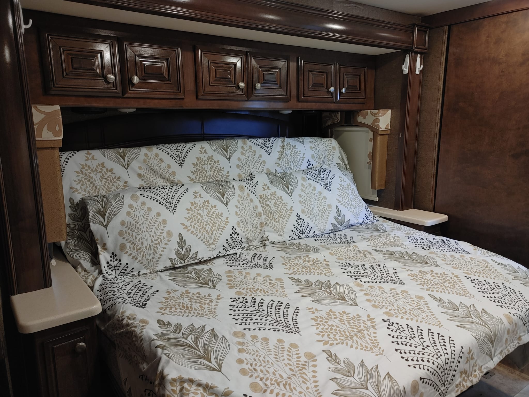 Detailed view of an RV rental bedroom with a bed, leaf-patterned duvet, dark wood cabinets, and side shelves.. Thor Palazzo 2017