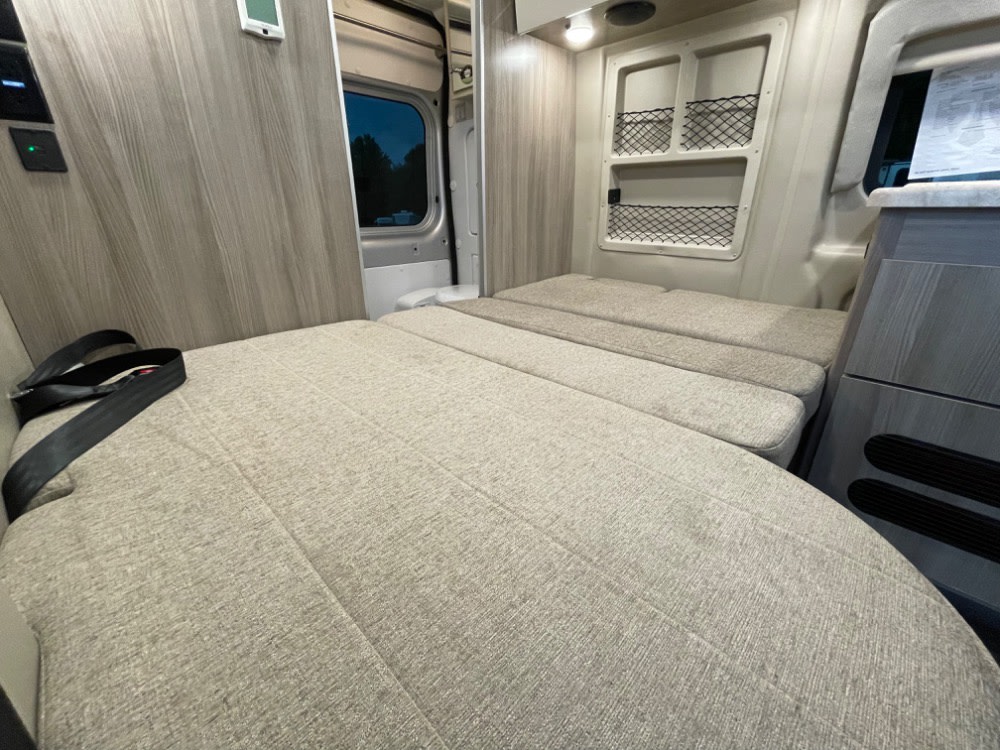 View of an RV rental interior, showing a bed/seating area made of light-colored cushions with a seatbelt. It's a compact living space.. Thor Scope 18G 2024