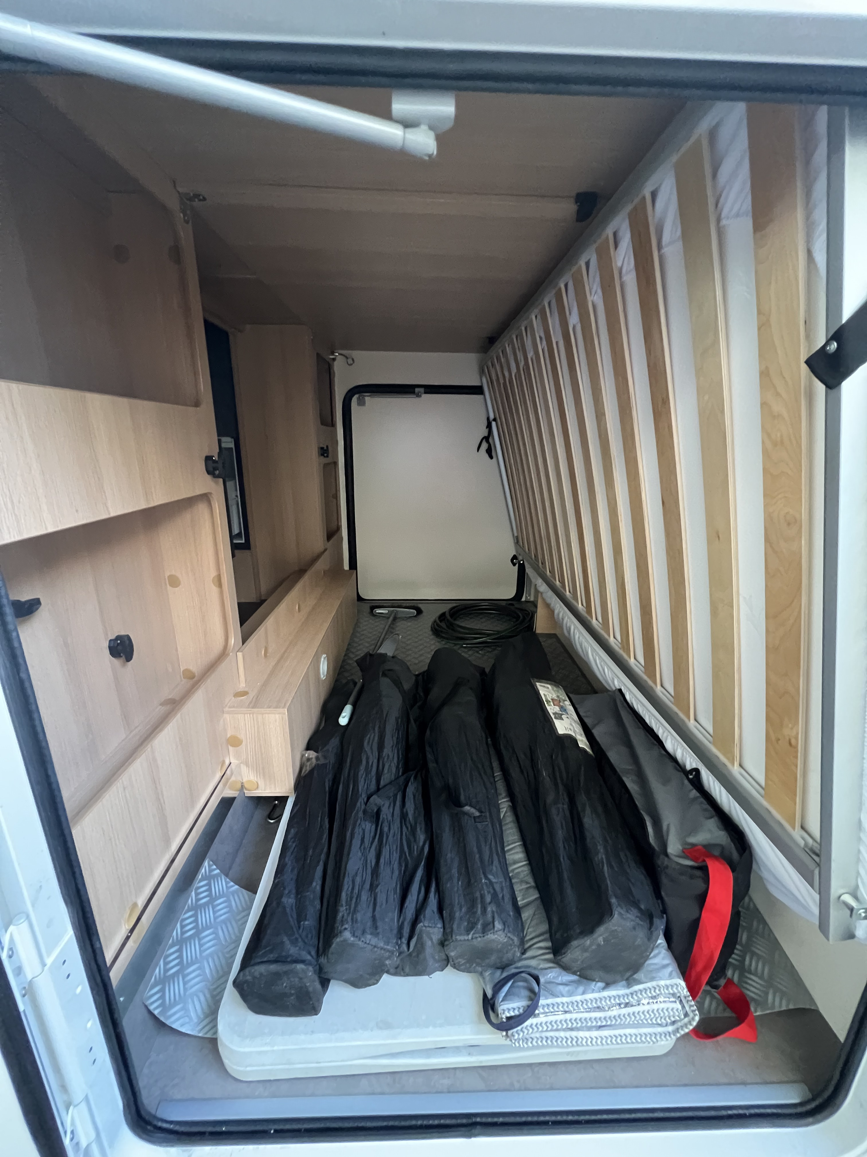 This RV rental interior features a large storage bay under a lifted slatted bed frame, holding black bags, a cable, and a small tool.. McLouis Glamys 2023