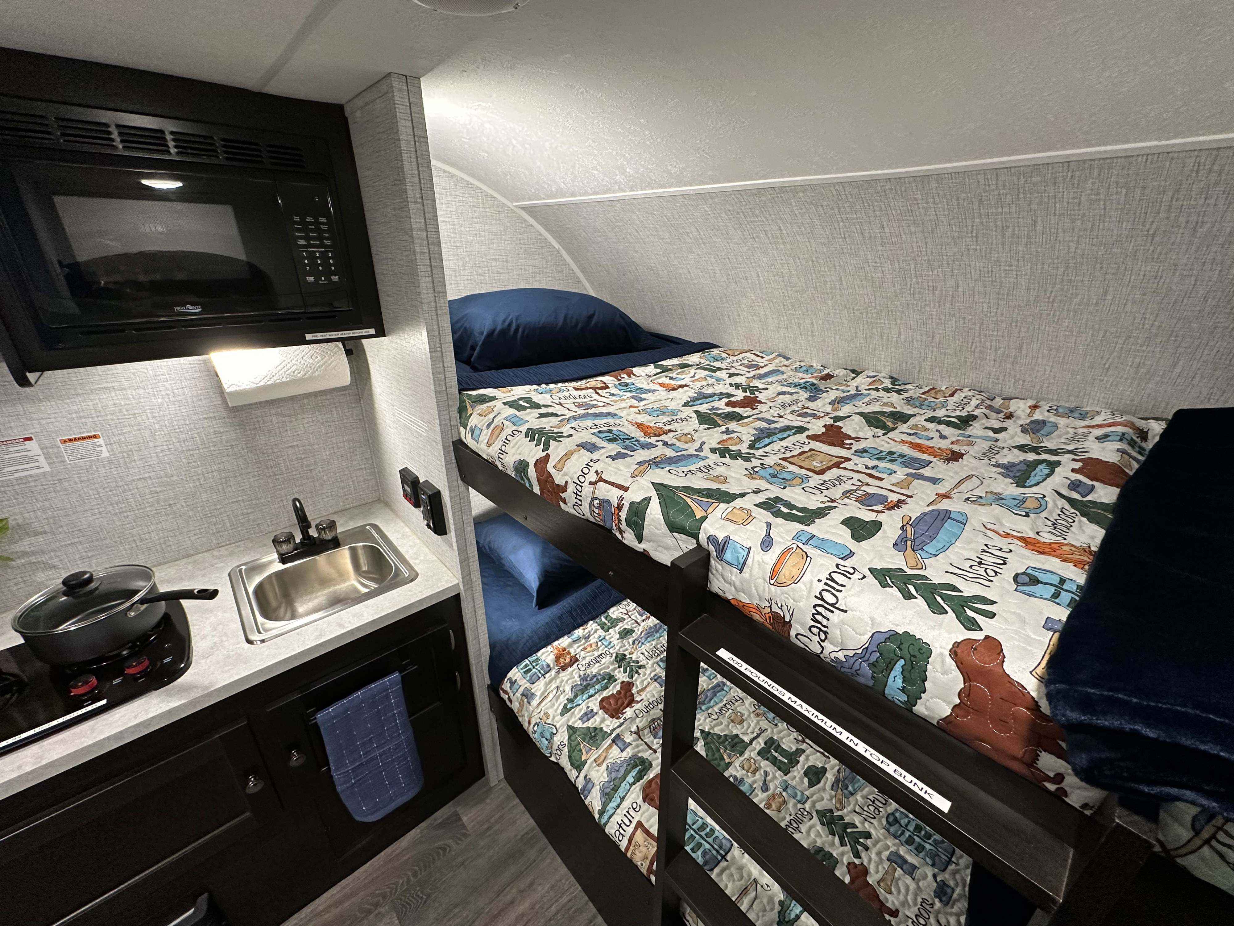 RV rental interior: a compact kitchen area with microwave, sink, and stovetop, adjacent to cozy bunk beds with themed quilts.. Braxton Creek Bushwhacker Plus 2022