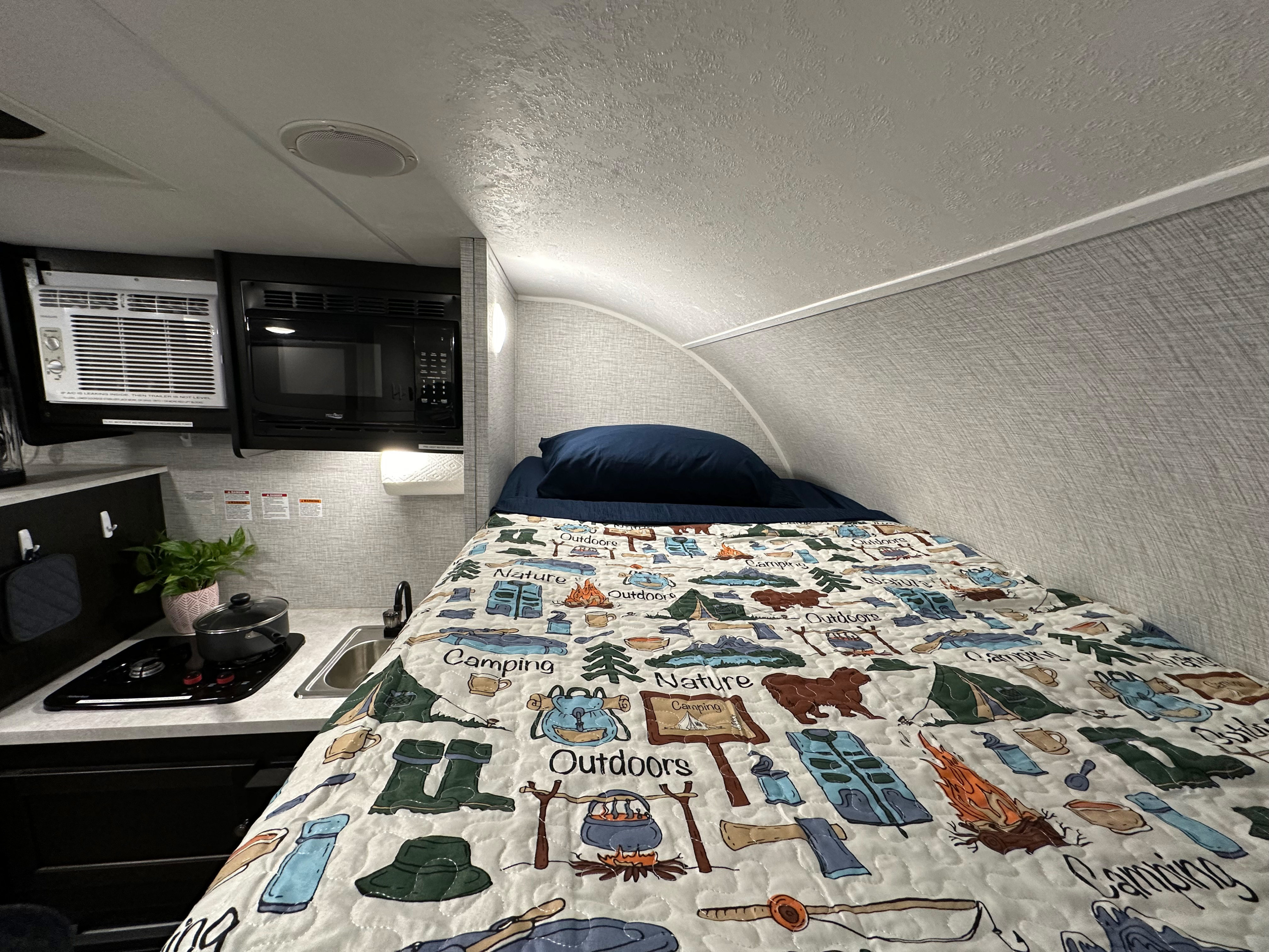Cozy RV rental interior: a compact space featuring a bed with a camping-themed quilt, kitchen, microwave, and AC unit.. Braxton Creek Bushwhacker Plus 2022