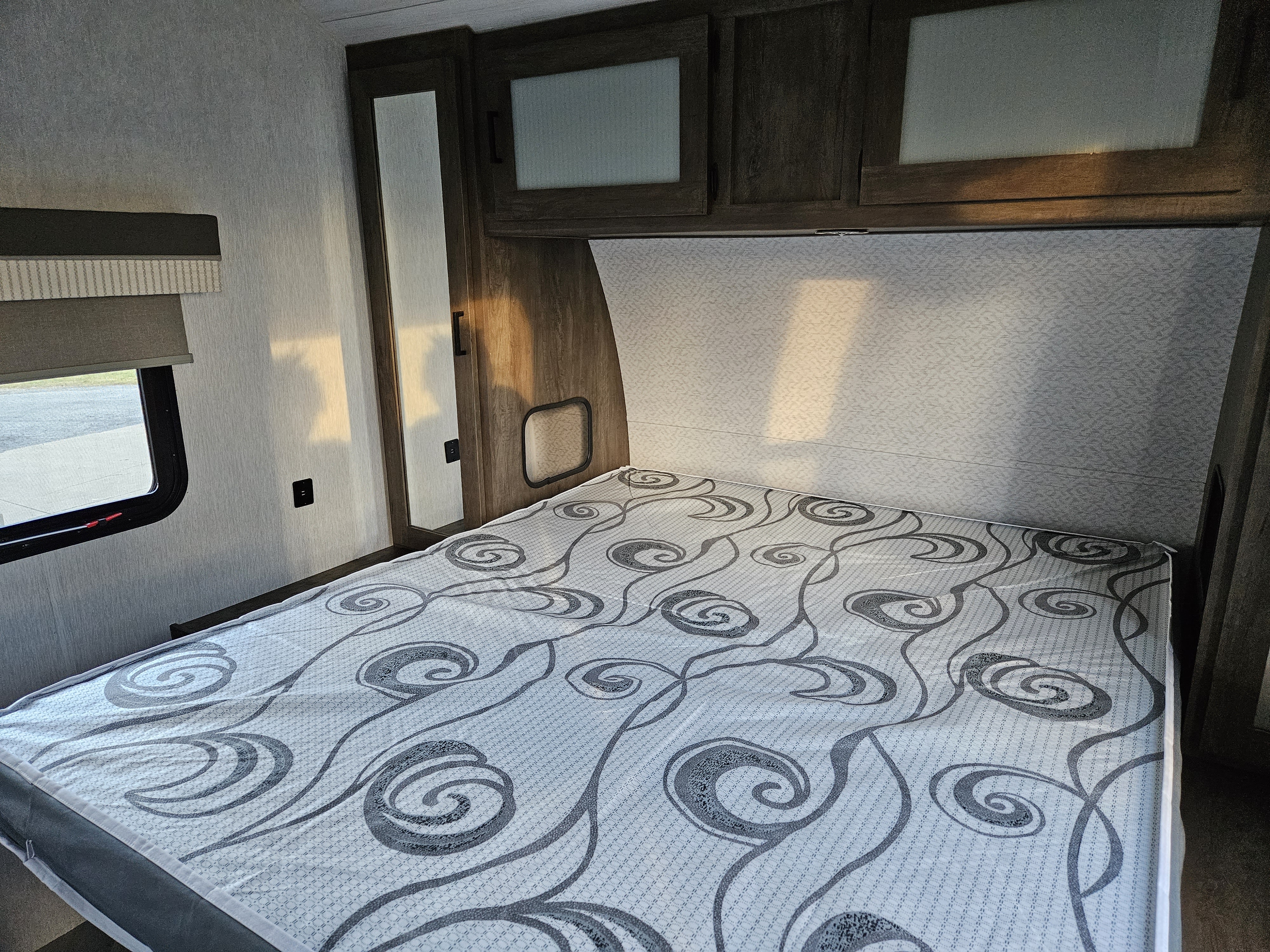 Modern RV rental bedroom. Features include a bed with patterned mattress, overhead cabinets, window with blind, and USB charging. Ideal for travel.. Forest River Wildwood 2022
