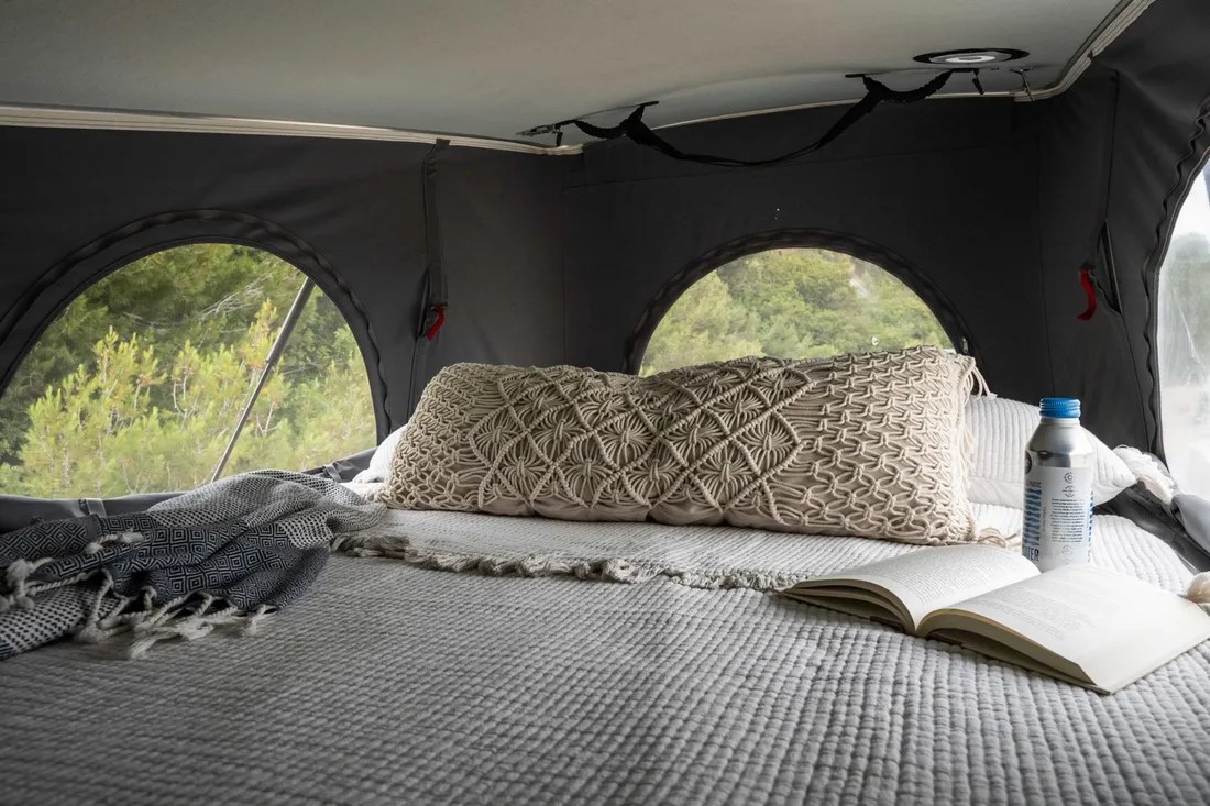 Inside a comfortable RV rental: a bed with an open book, water bottle, and scenic forest views from arched windows.. Winnebago Solis 2022