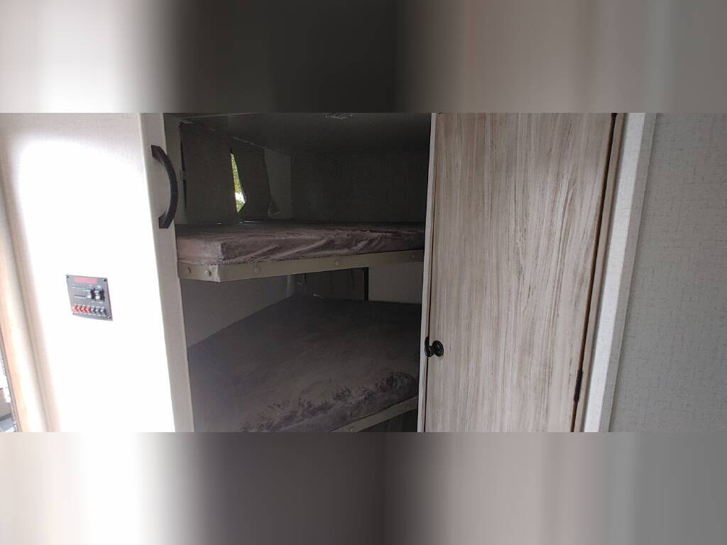 Inside an RV rental: two bunk beds with mattresses, a control panel on the wall, and a light-colored wood door, ideal for travel.. Winnebago 2301BHS 2022
