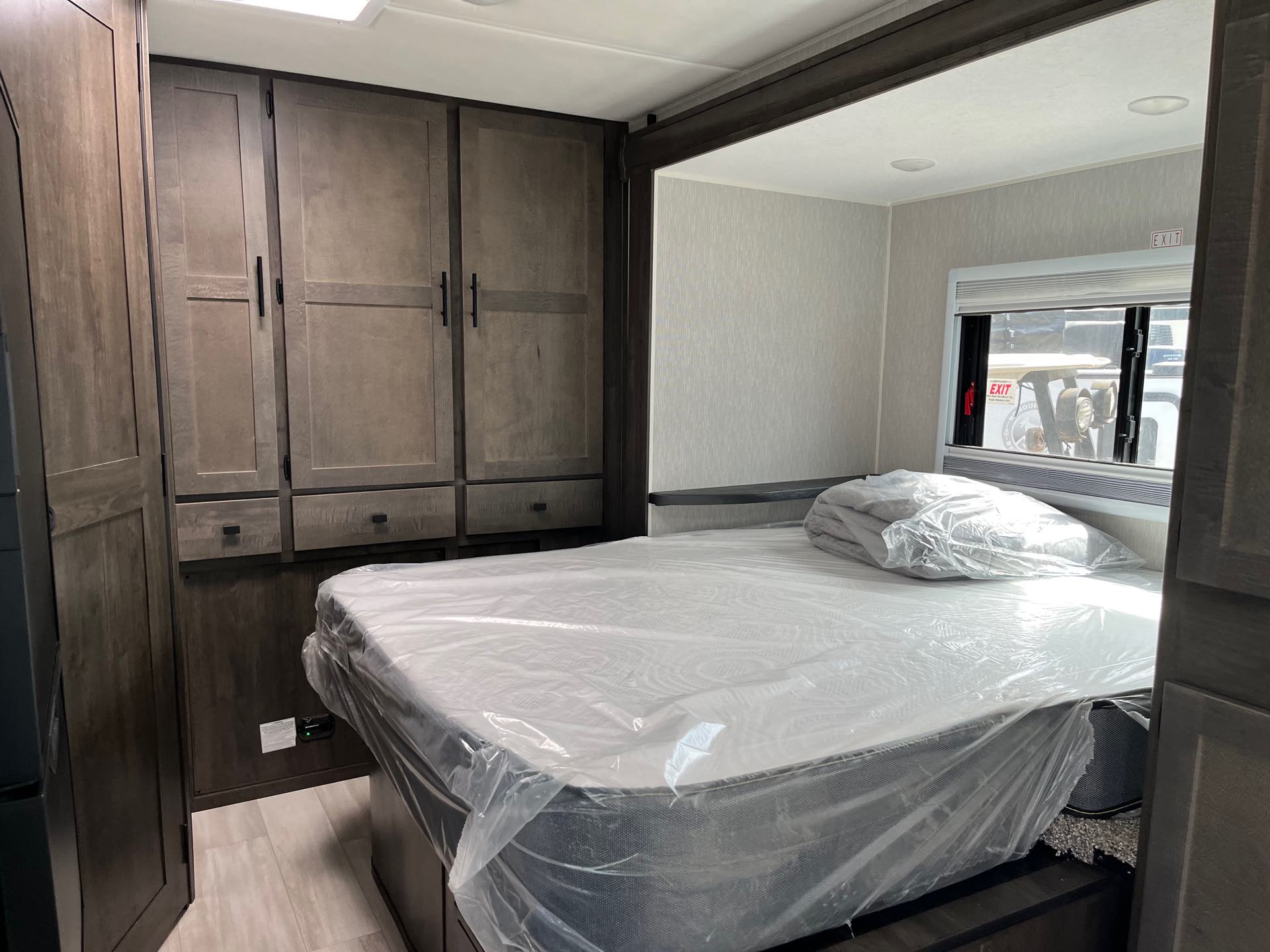 Inside a modern RV rental bedroom: a bed with a protective cover, ample dark wood cabinets, and a window with blinds.. Coachmen Prism Elite 2024