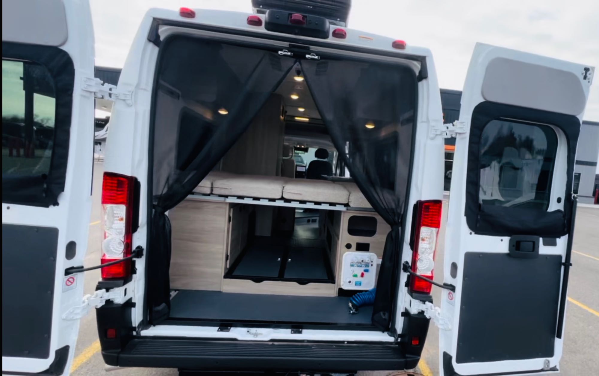 Rear of a white camper van RV rental, doors open to reveal an elevated bed, under-bed storage, and a protective mesh screen.. Winnebago Solis 59px 2023