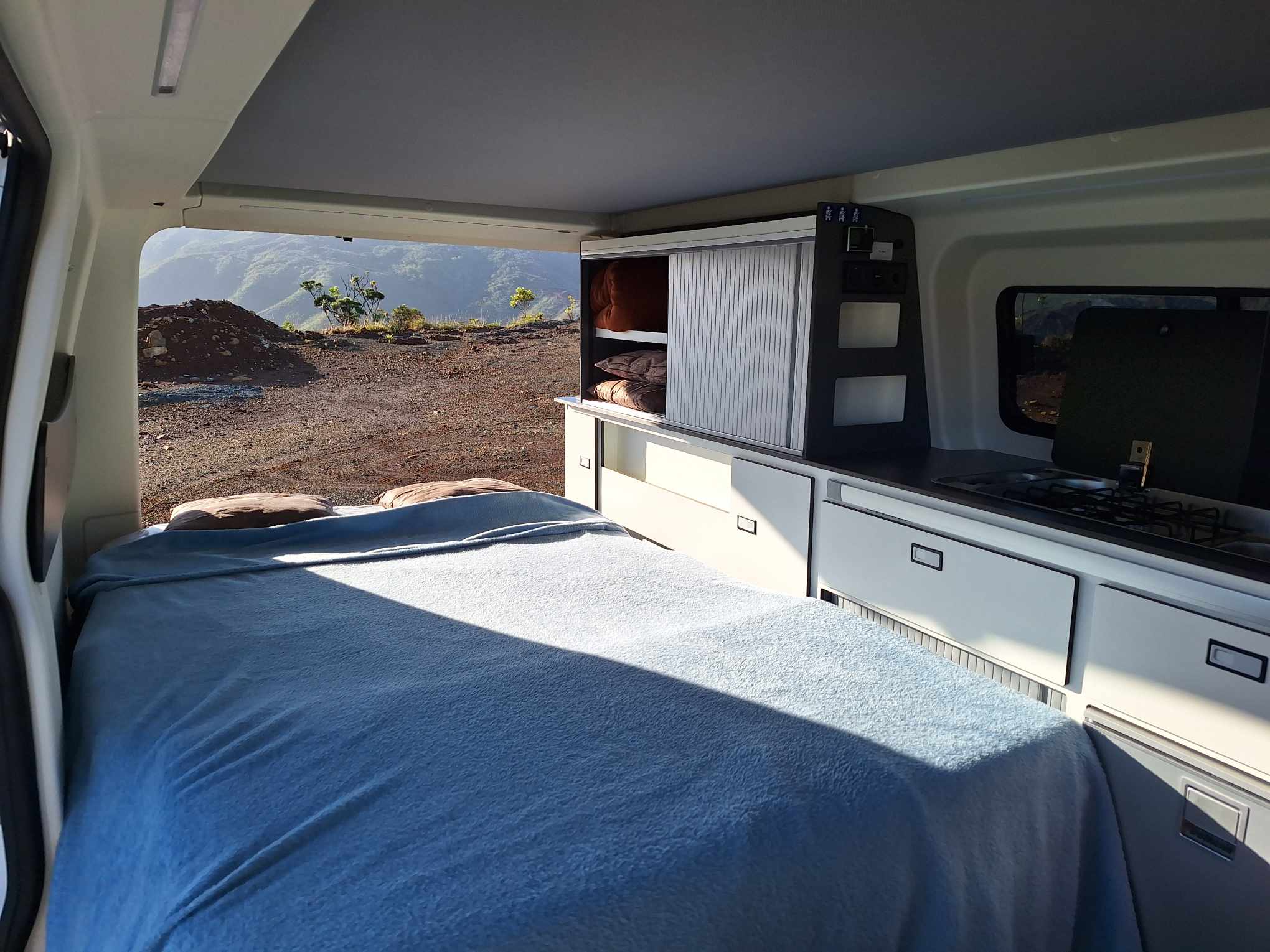 An rv rental camper van interior shows a blue bed, kitchen, and open rear door overlooking a mountain landscape.. Panama P10+ 2024