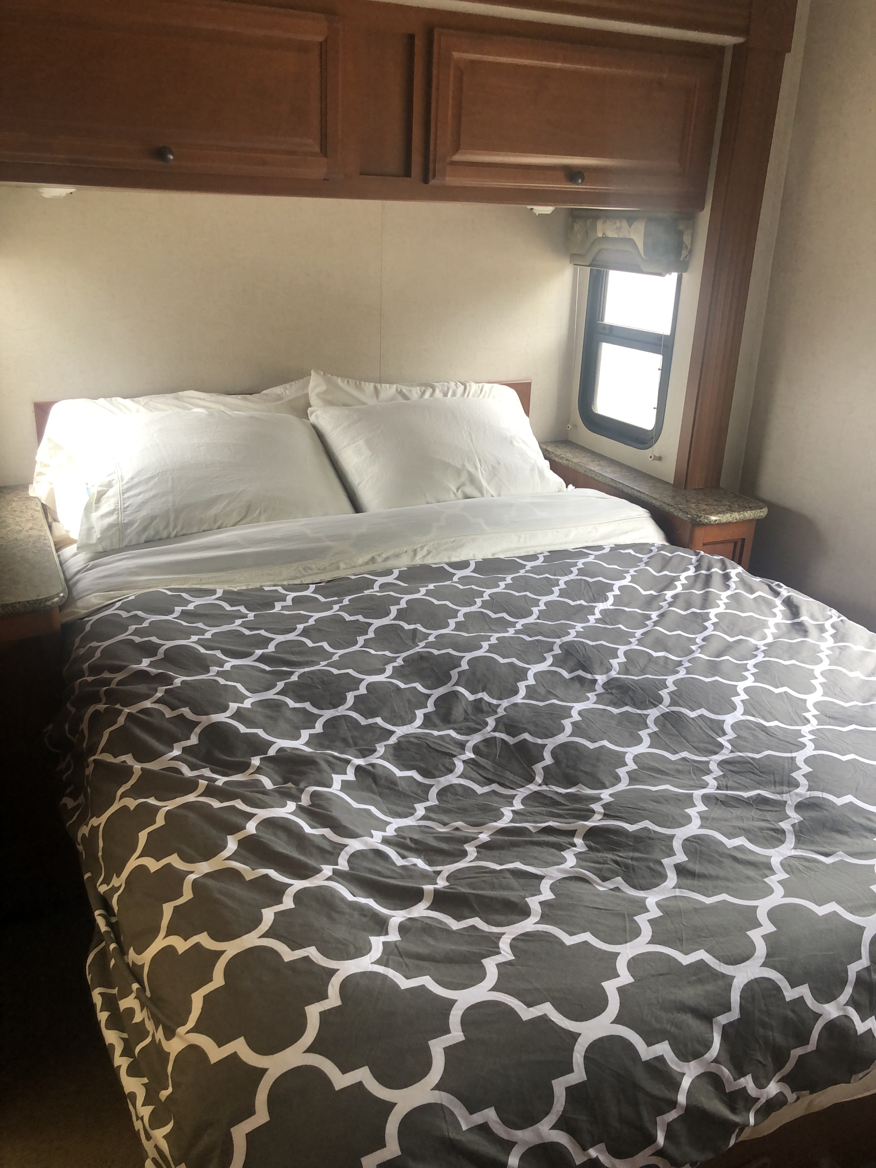 A cozy RV rental bedroom features a bed with white pillows, grey patterned comforter, overhead cabinets & side window.. Jamboree Sport 2010