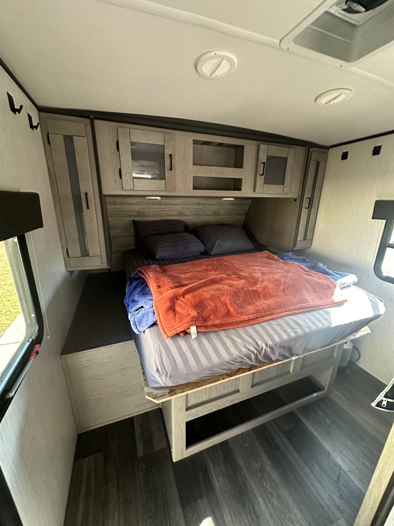 RV rental bedroom: striped bed with orange blanket, dark pillows, wood-look floor, and light wood overhead cabinets.. Heartland RVs Mallard 2021