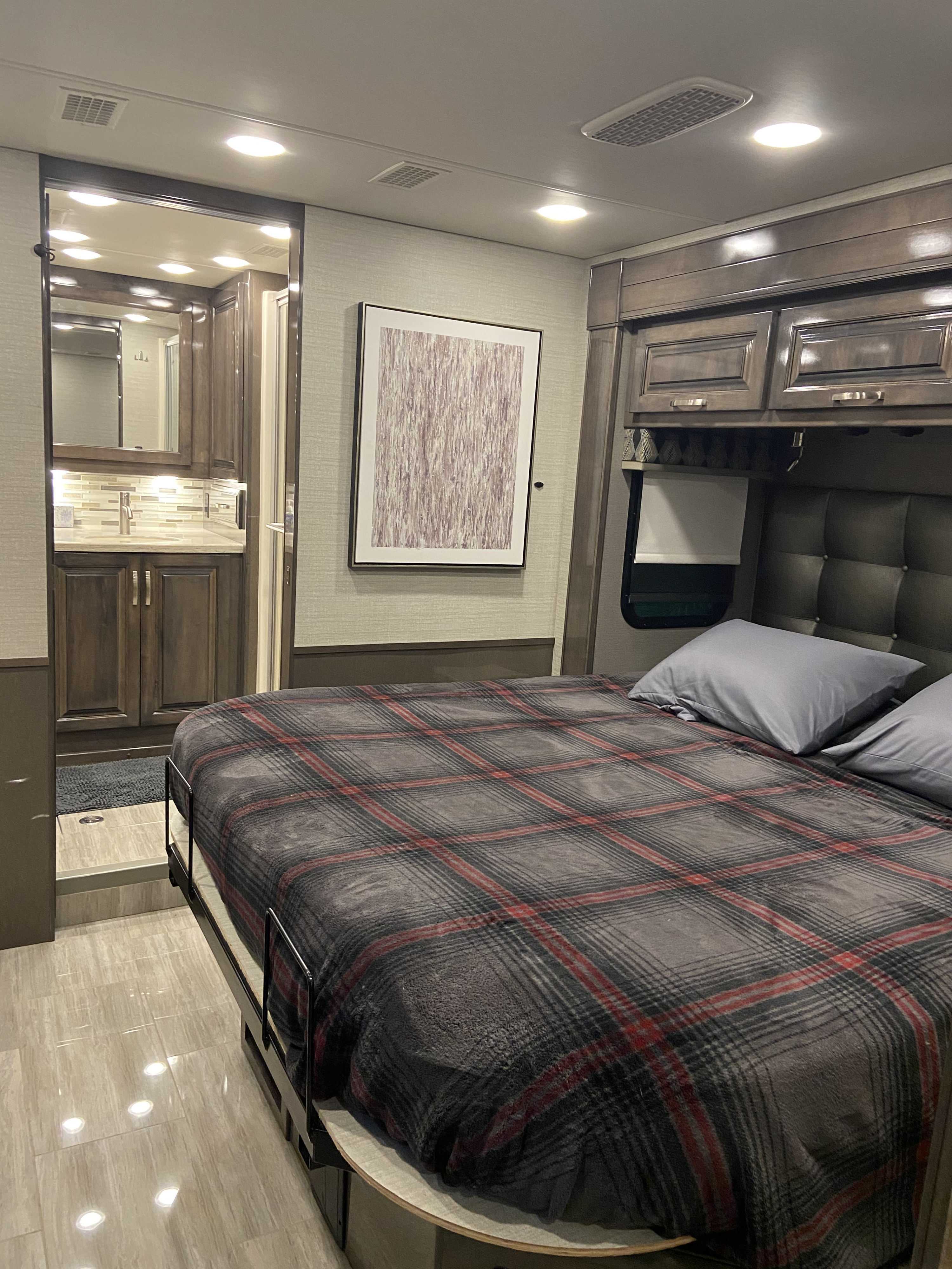 Cozy RV rental interior showing a bedroom with a plaid blanket and an adjacent bathroom with a vanity.. Entegra Coach Reatta XL 2020