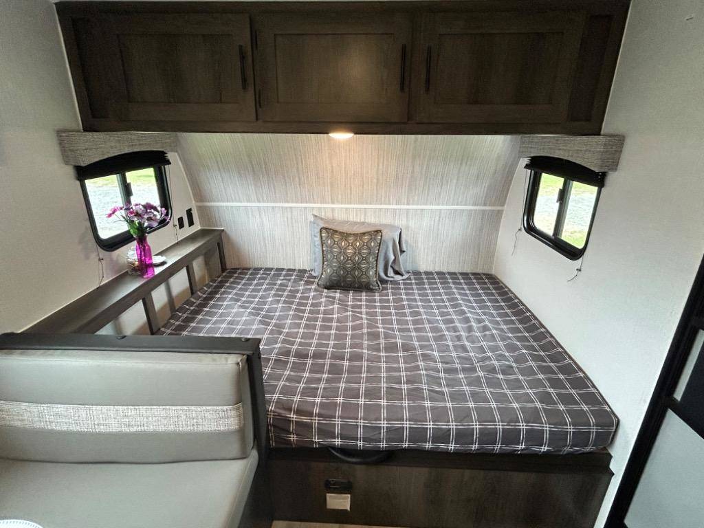Inside an RV rental: a compact bedroom with a plaid bed, overhead cabinets, and dual windows. Cozy travel space.. Coachmen Clipper 2024