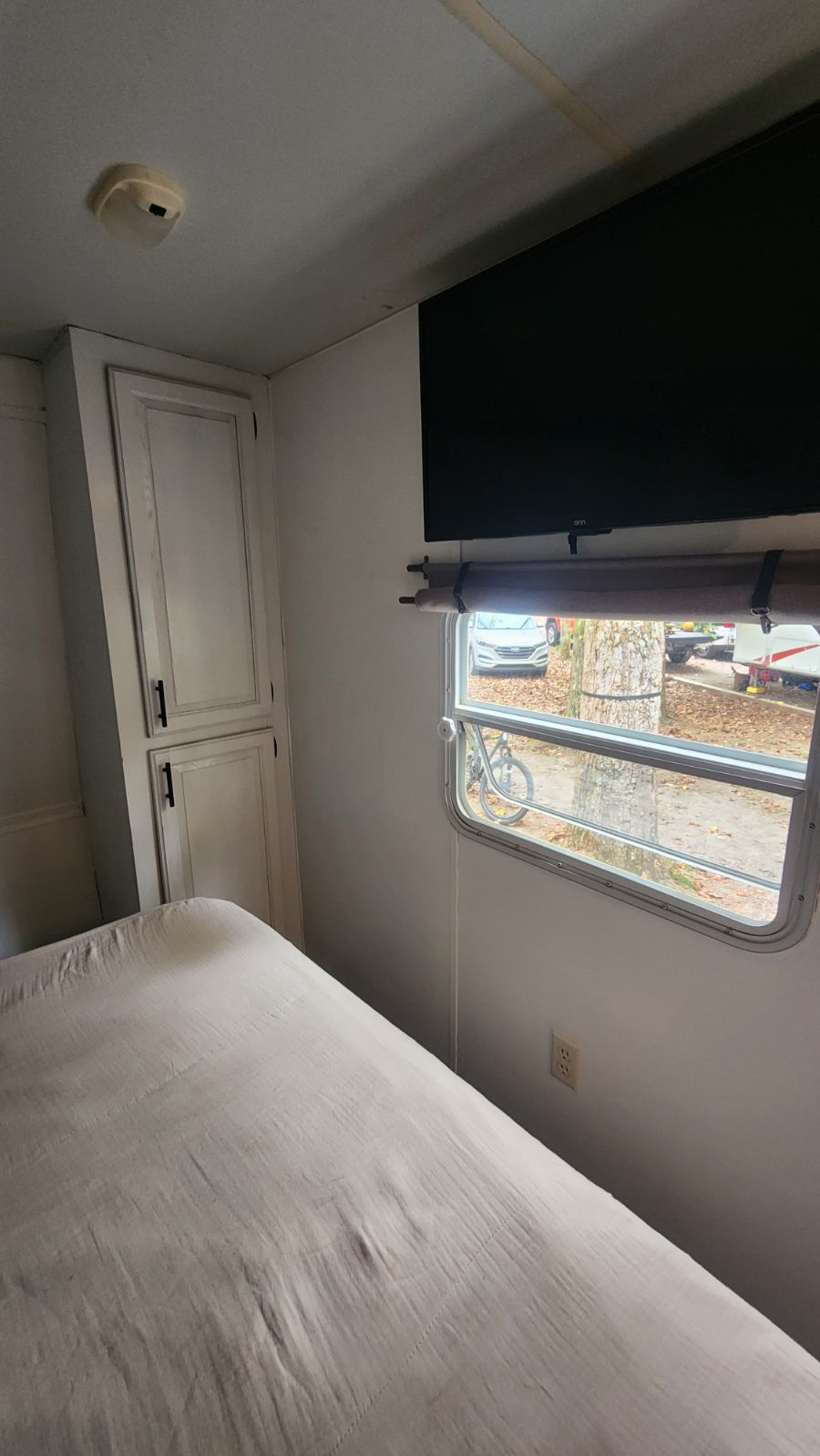 Cozy RV rental bedroom with a bed, white cabinet, wall-mounted TV, and window overlooking a leafy campground.. Timberland Timberlodge 2003