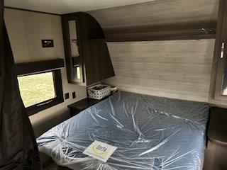 Step into this comfortable RV rental bedroom, featuring a new bed with plastic covering, nightstand, and window for your next travel adventure.. Jayco Jay Flight SLX 2021