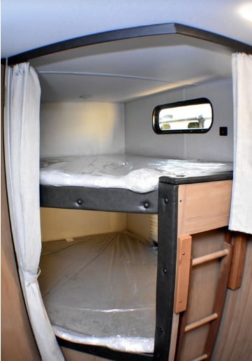 Inside an RV rental, two bunk beds with black frames and plastic-wrapped mattresses are visible. An oval window and ladder complete the setup.. Forest River Cherokee Grey Wolf 2023