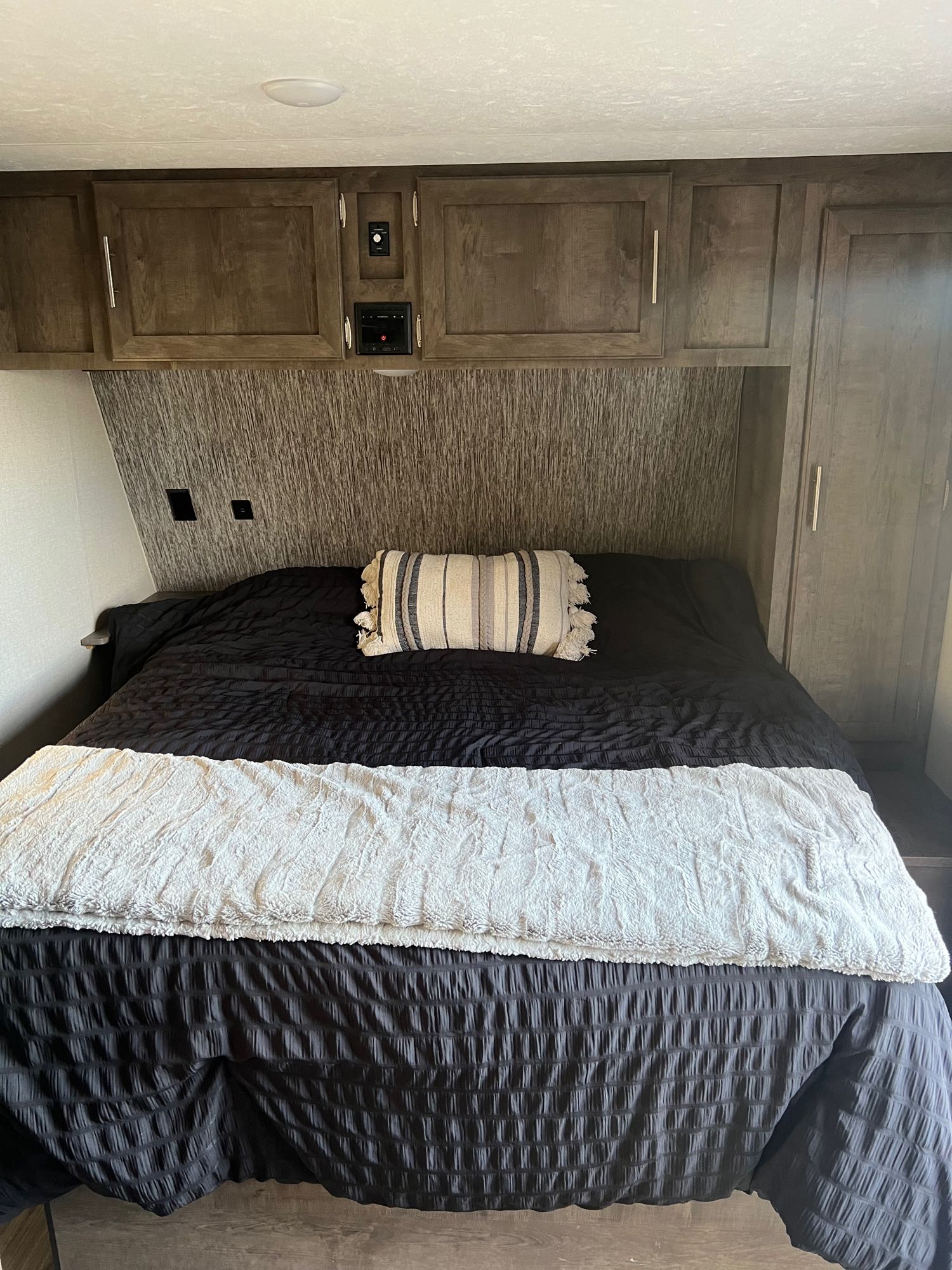 Cozy RV rental bedroom featuring a bed with dark bedding, striped pillow, and wood overhead cabinets for storage.. KZ Escape 2019