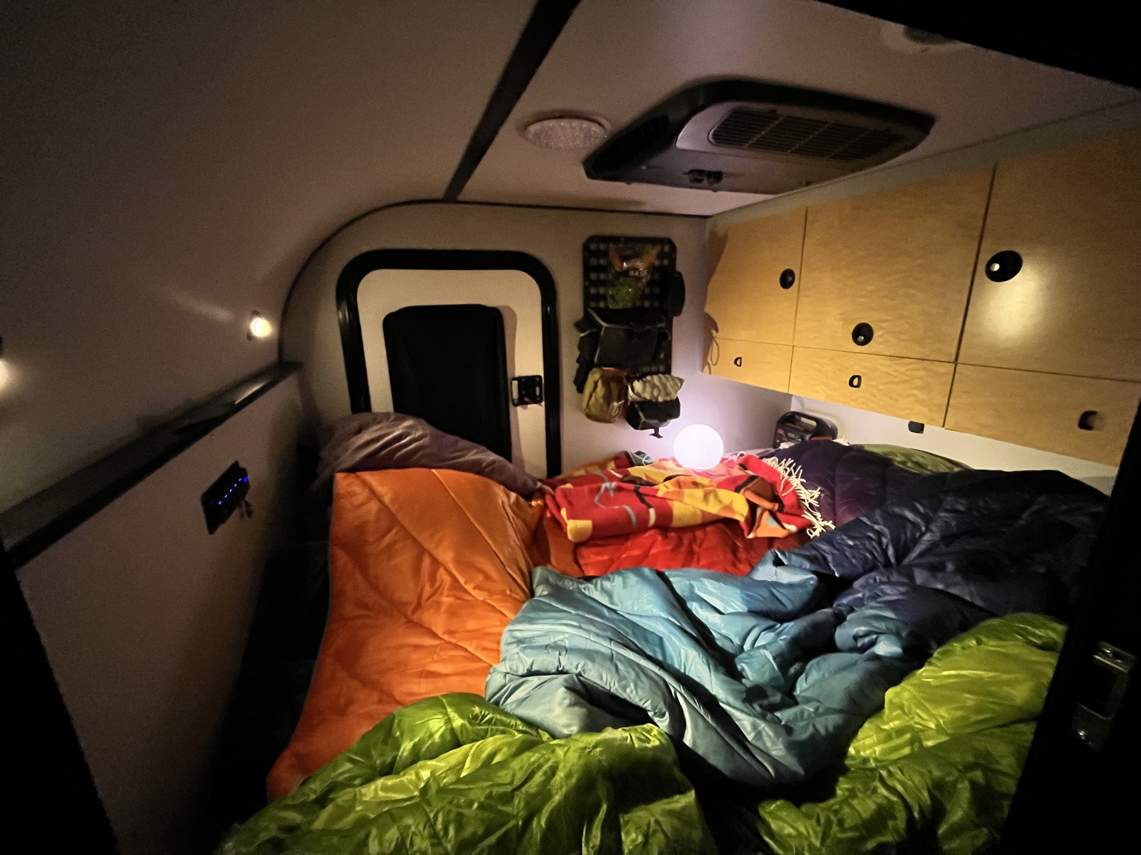 Cozy RV rental interior at night, showing multiple colorful sleeping bags, a glowing spherical lamp, and overhead wooden cabinets.. BRX Overland trailer 2021