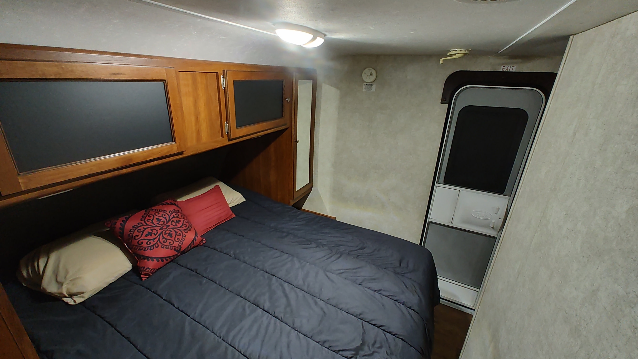 Inside an RV rental, a comfy bedroom features a dark bed, wooden overhead cabinets, and an exit door, ensuring cozy travels.. Keystone RV Bullet Ultra Lite 2015
