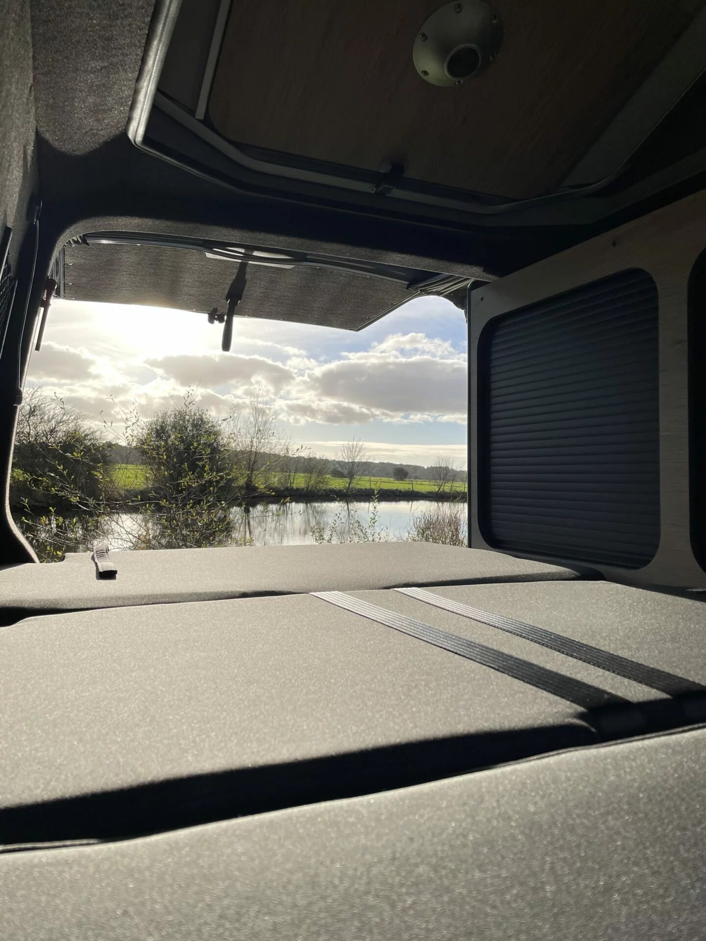 Cozy RV rental interior with bed looking out to a serene lake, green fields, and bright sky with clouds.. Volkswagen Summer 2024