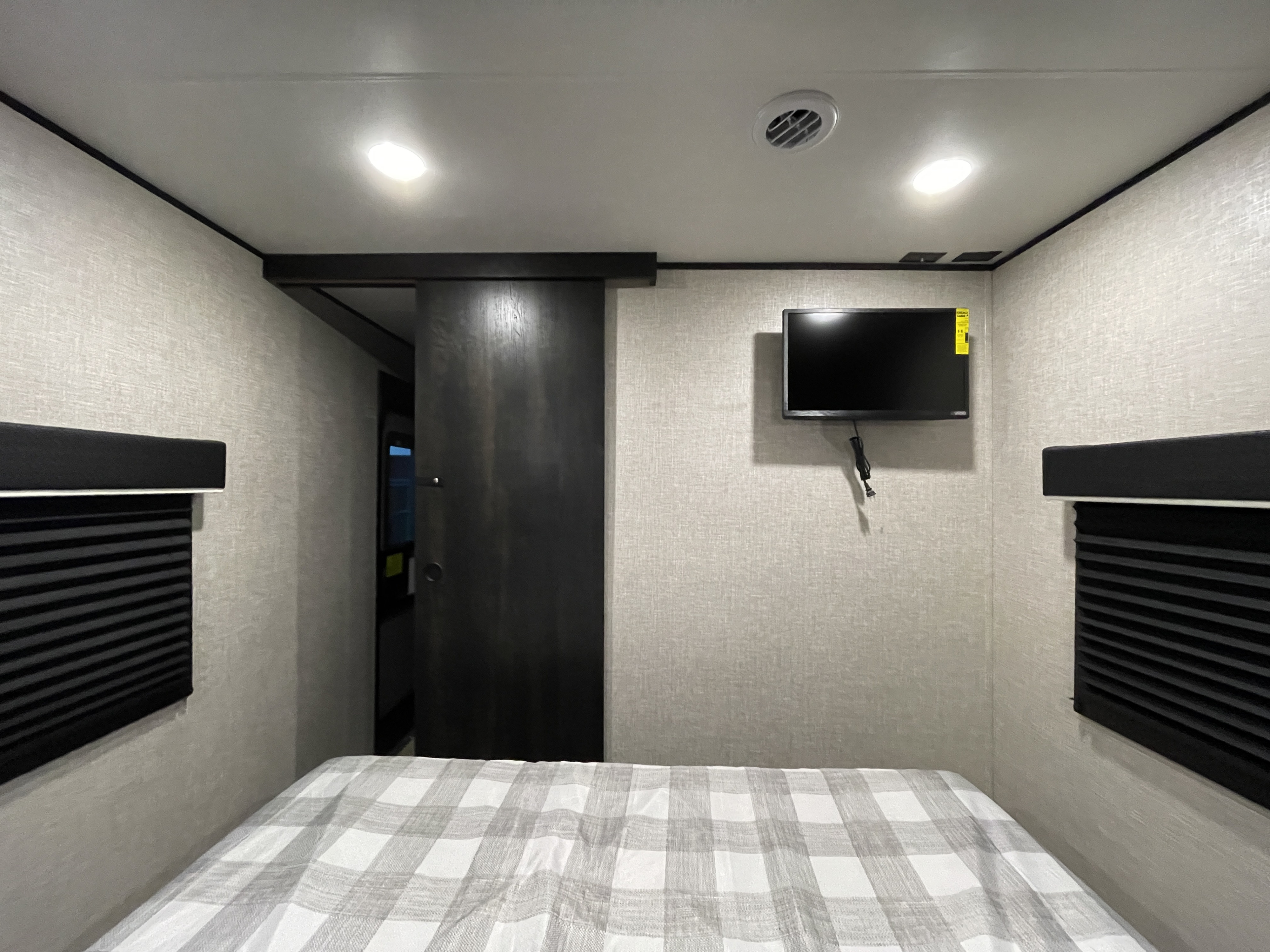 This RV rental bedroom features a comfy plaid bed, wall-mounted TV, and window blinds, with a sliding door on the left.. Jayco Jay Flight SLX 2022