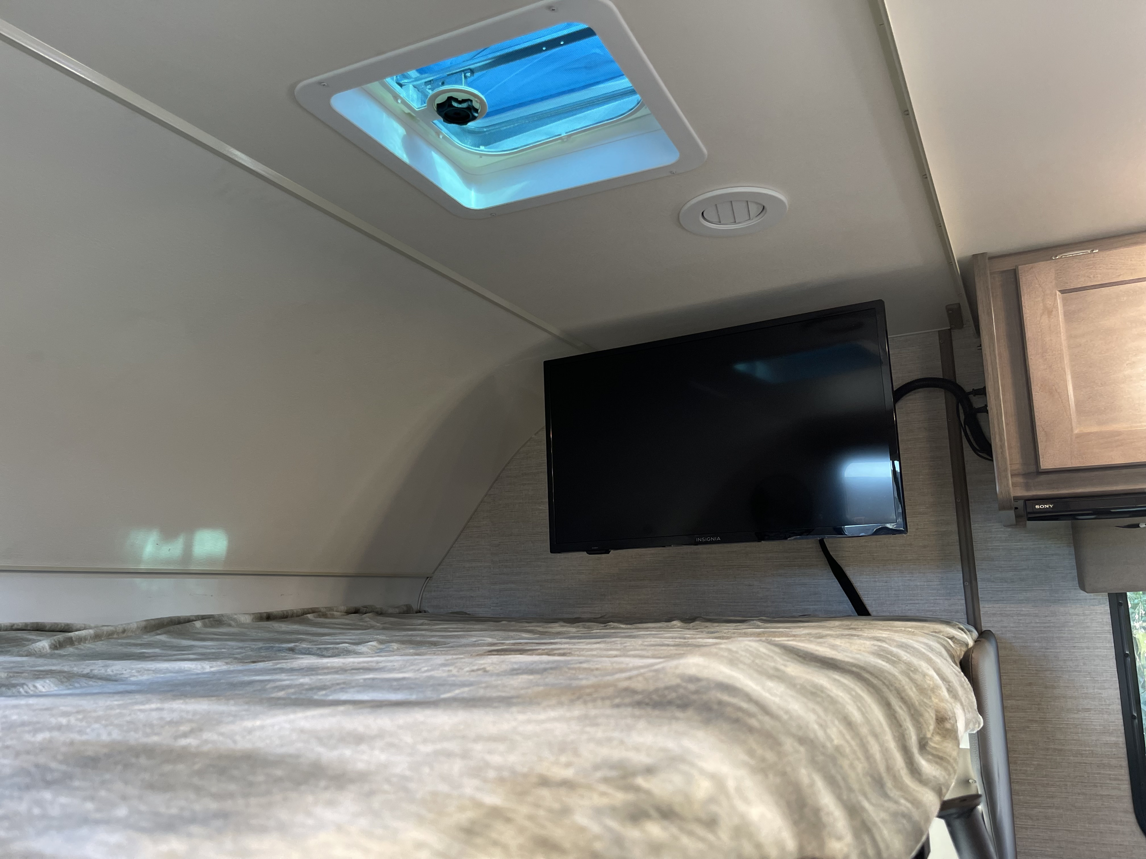 Inside an RV rental: a cozy bunk with a mounted TV, overhead vent, and bright skylight.. Winnebago Minnie Winnie 2024