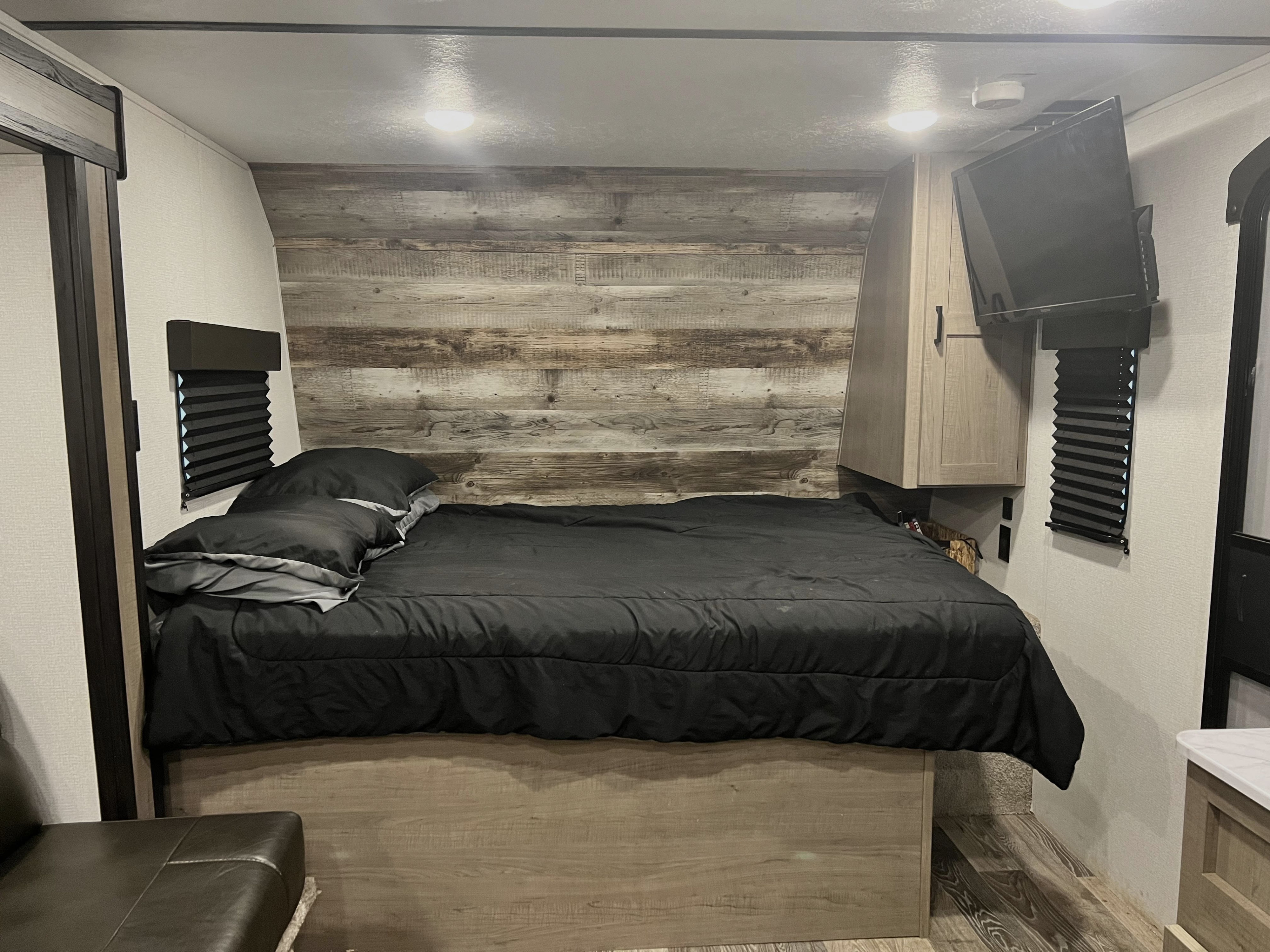 Cozy RV rental bedroom featuring a bed with black bedding, wood accent wall, mounted TV, and small windows.. Keystone RV Hideout 2021