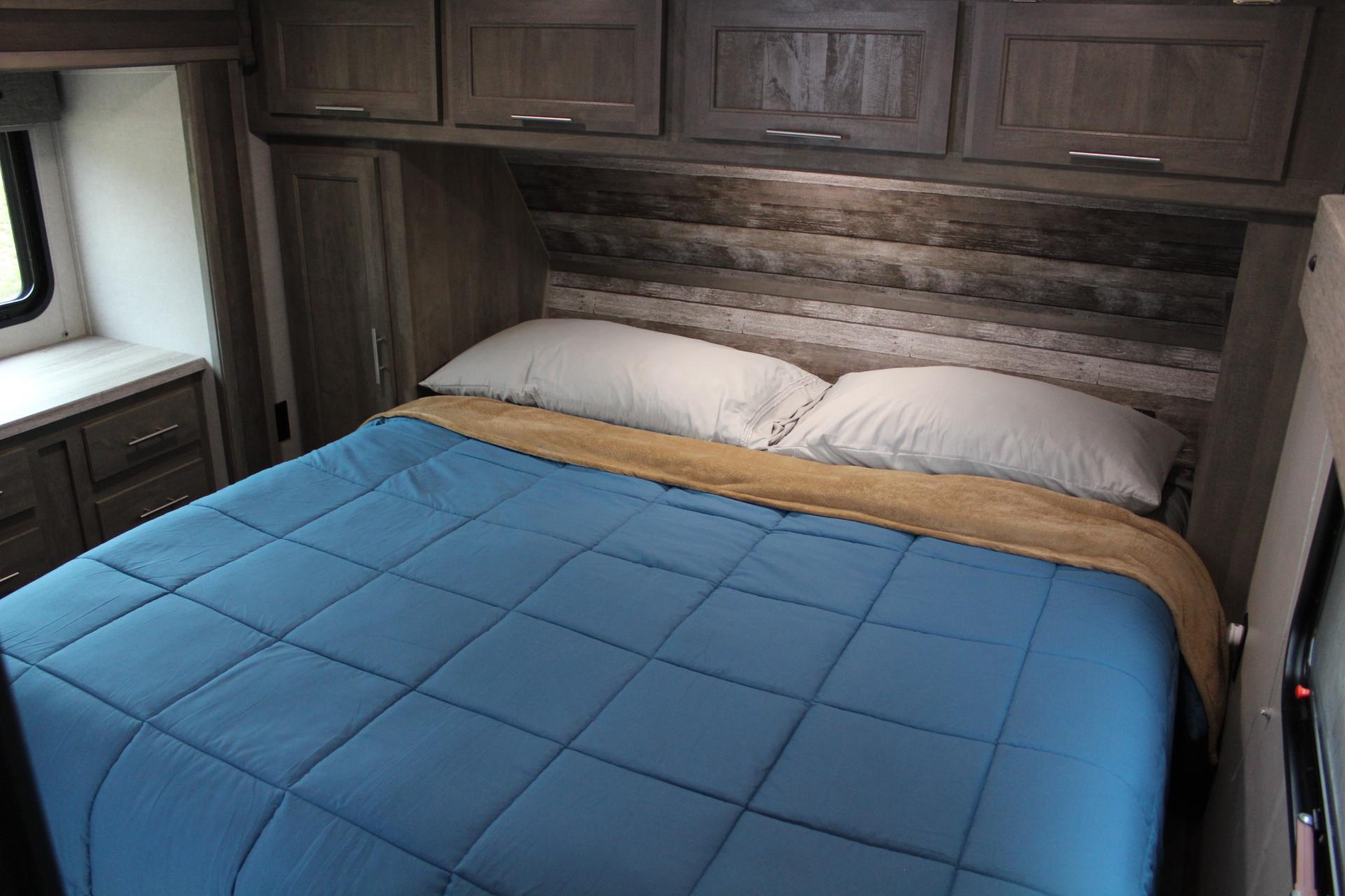 RV rental bedroom: comfortable bed with blue comforter, rustic wood headboard, overhead cabinets & window.. Forest River XLR Boost Toy Hauler 2021