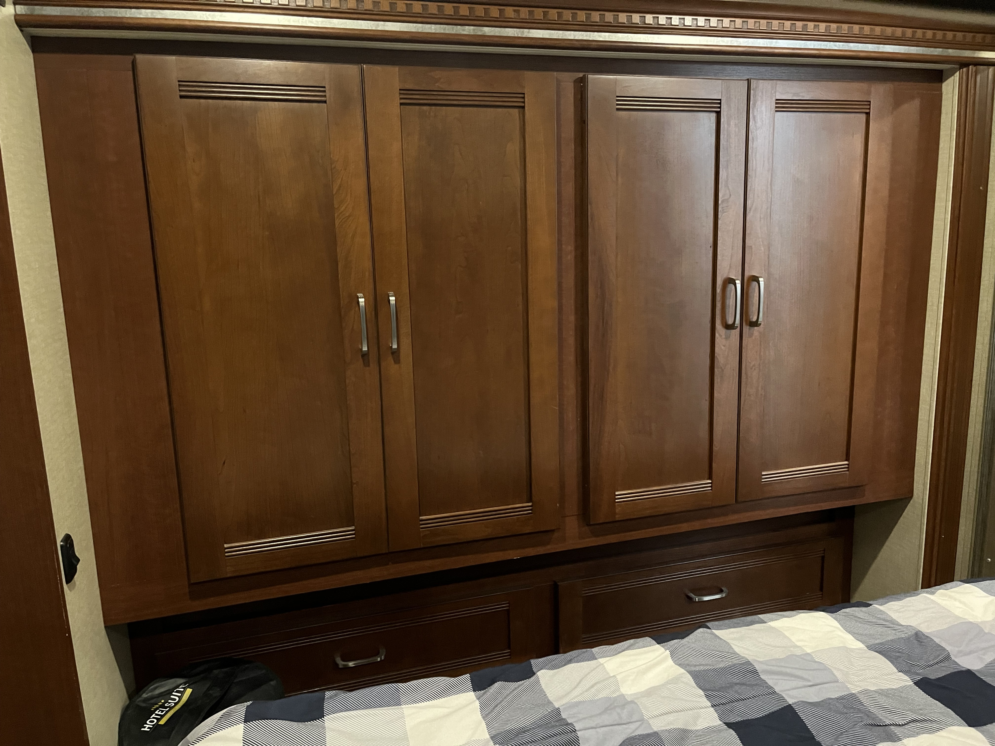 Inside an RV rental: dark wood overhead storage cabinets, handles, and lower drawers above a plaid bed. Compact bedroom space.. Winnebago Forza 2014