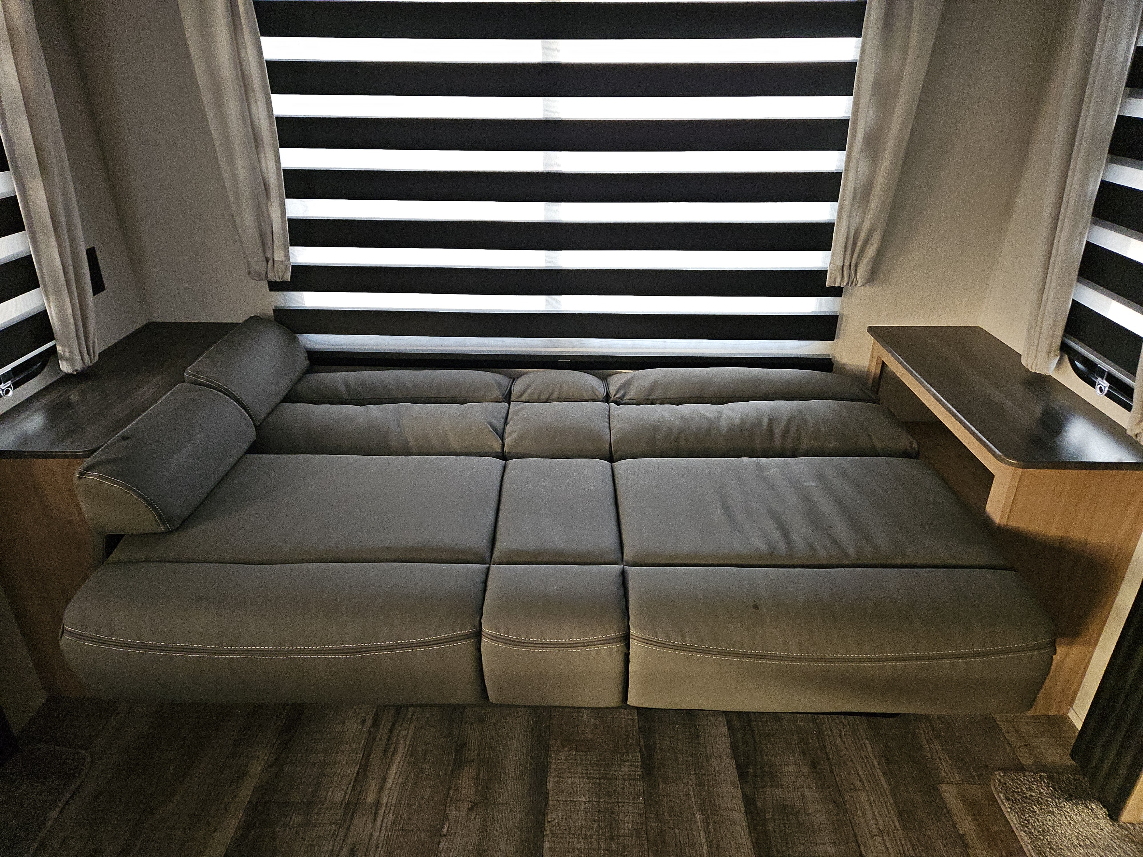 RV rental interior showcasing a grey convertible sofa, wood floor, side tables, and striped window blinds.. Forest River Cherokee Grey Wolf 2023