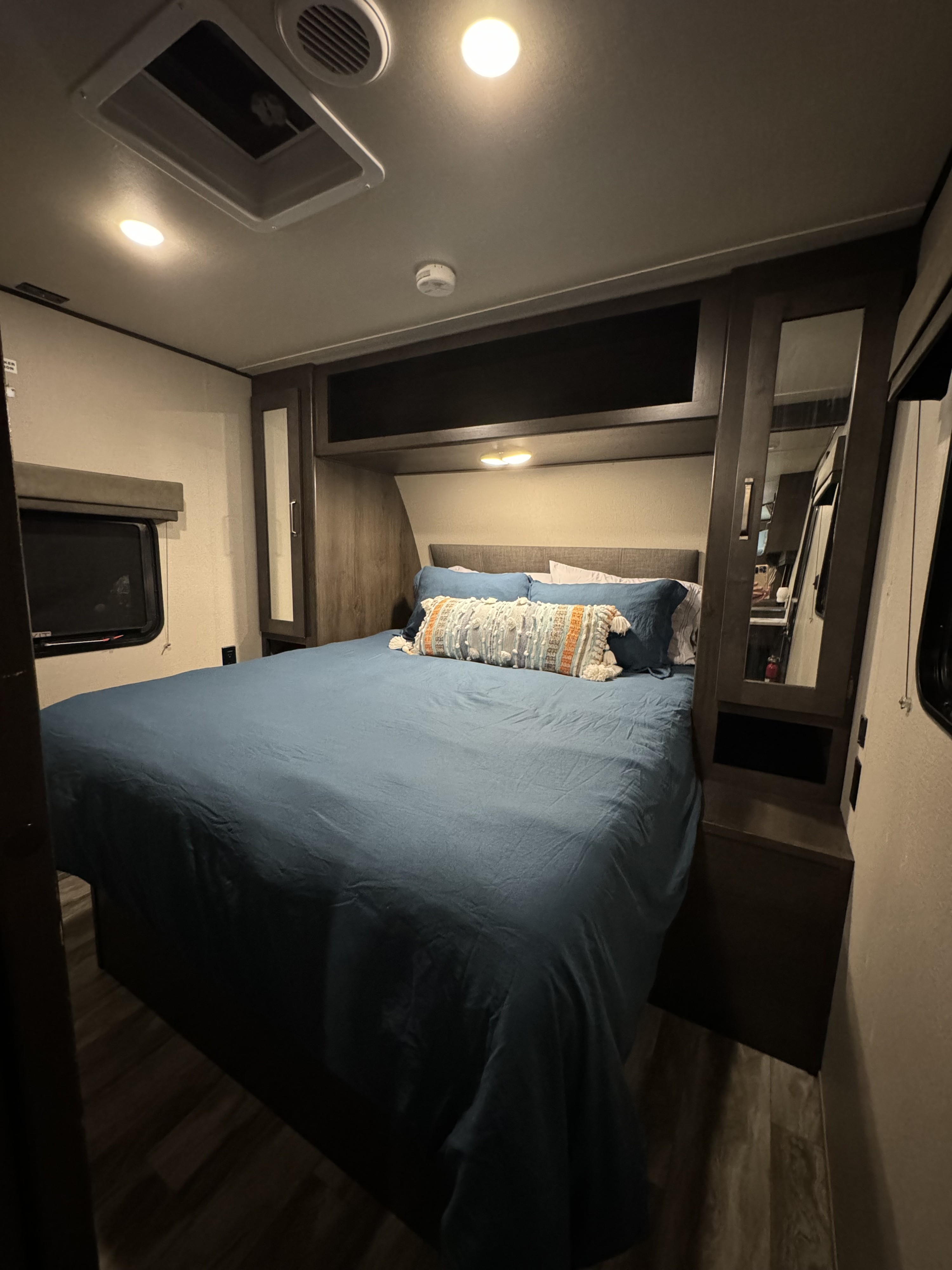 This inviting RV rental bedroom features a queen-sized bed with blue bedding, overhead storage, and a window, offering comfort on the go.. Grand Design Transcend Xplor 2021