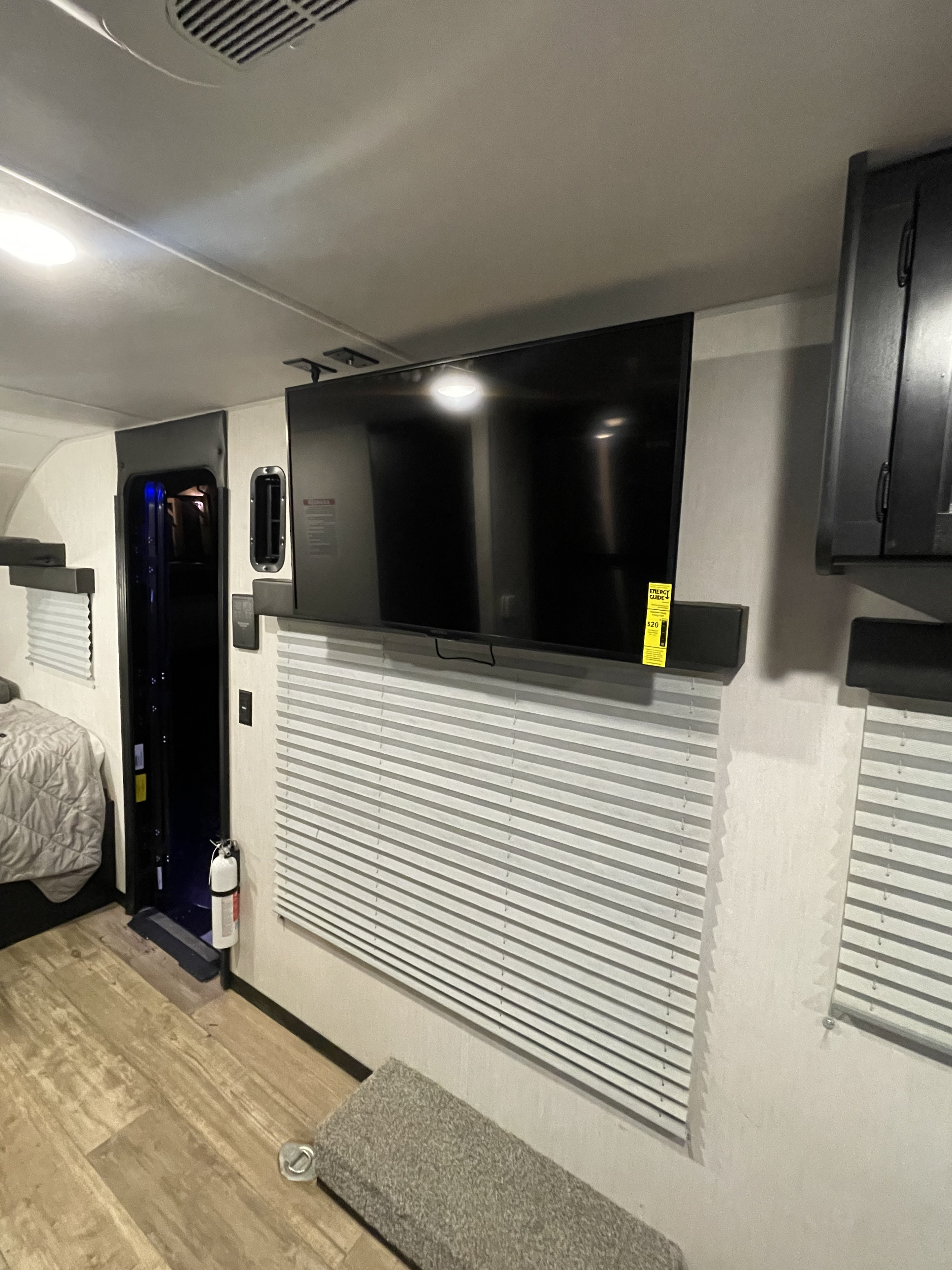 RV rental interior view: large wall-mounted TV, soundbar, and pleated blinds. Includes a bed area, exit door, and safety features.. Forest River Wolf Pup 2022