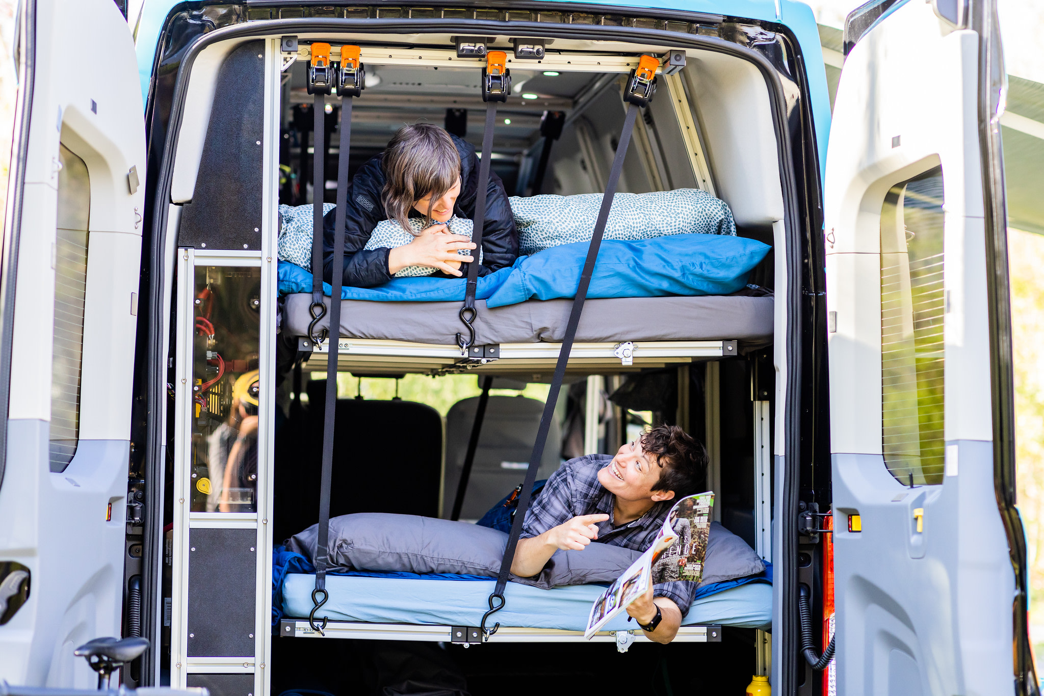 Two people relaxing on bunk beds inside an RV rental van, seen through its open rear doors. Perfect for camping.. Ford Transit 350 HD XLT 2019